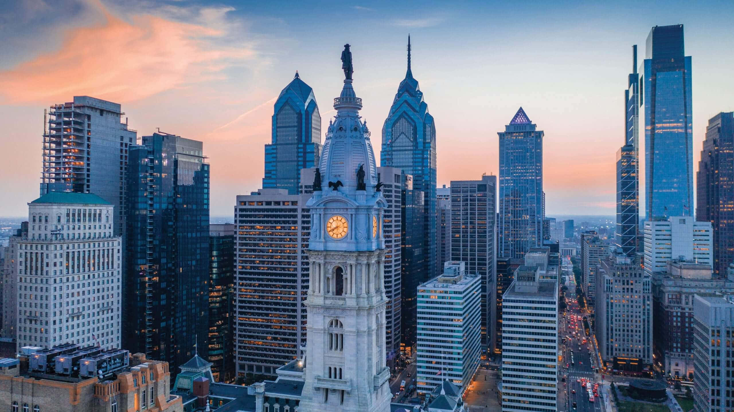 Philly 桌布 壁紙 — 費城天際線在日落時的壯麗鳥瞰圖，標誌性的市政廳鐘樓在現代玻璃摩天大樓的映襯下溫暖地發光。