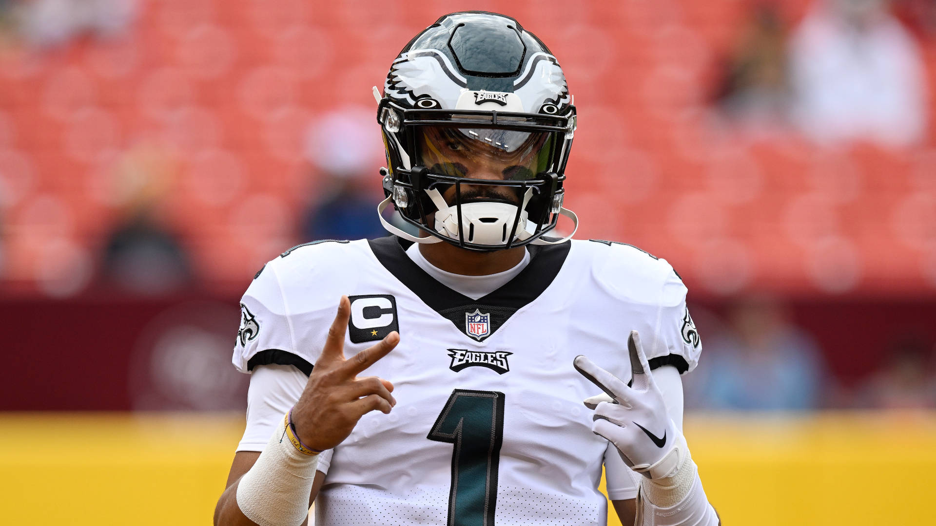 Jalen Hurts Eagles 桌布 壁紙 — 費城老鷹隊四分衛穿著白色球衣，帶有隊長'C'和1號，在球場上做出和平手勢。