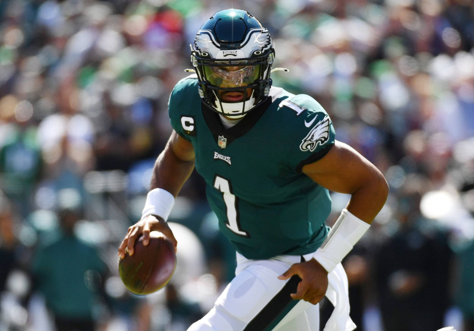 Jalen Hurts Eagles 桌布 壁紙 — 費城老鷹隊四分衛穿著綠色球衣，在日間比賽中帶著橄欖球奔跑，背景是模糊的人群。