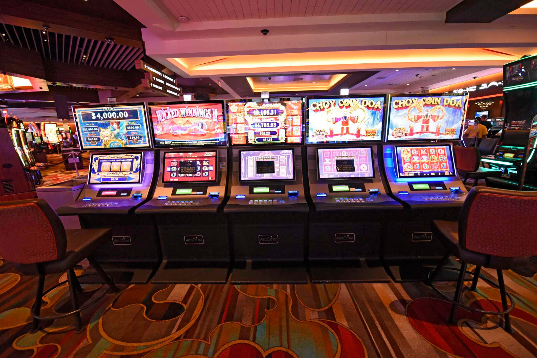 game 桌布 壁紙 — 賭場內一排明亮照明的老虎機，配有彩色螢幕和圖案地毯，在閃爍燈光下。
