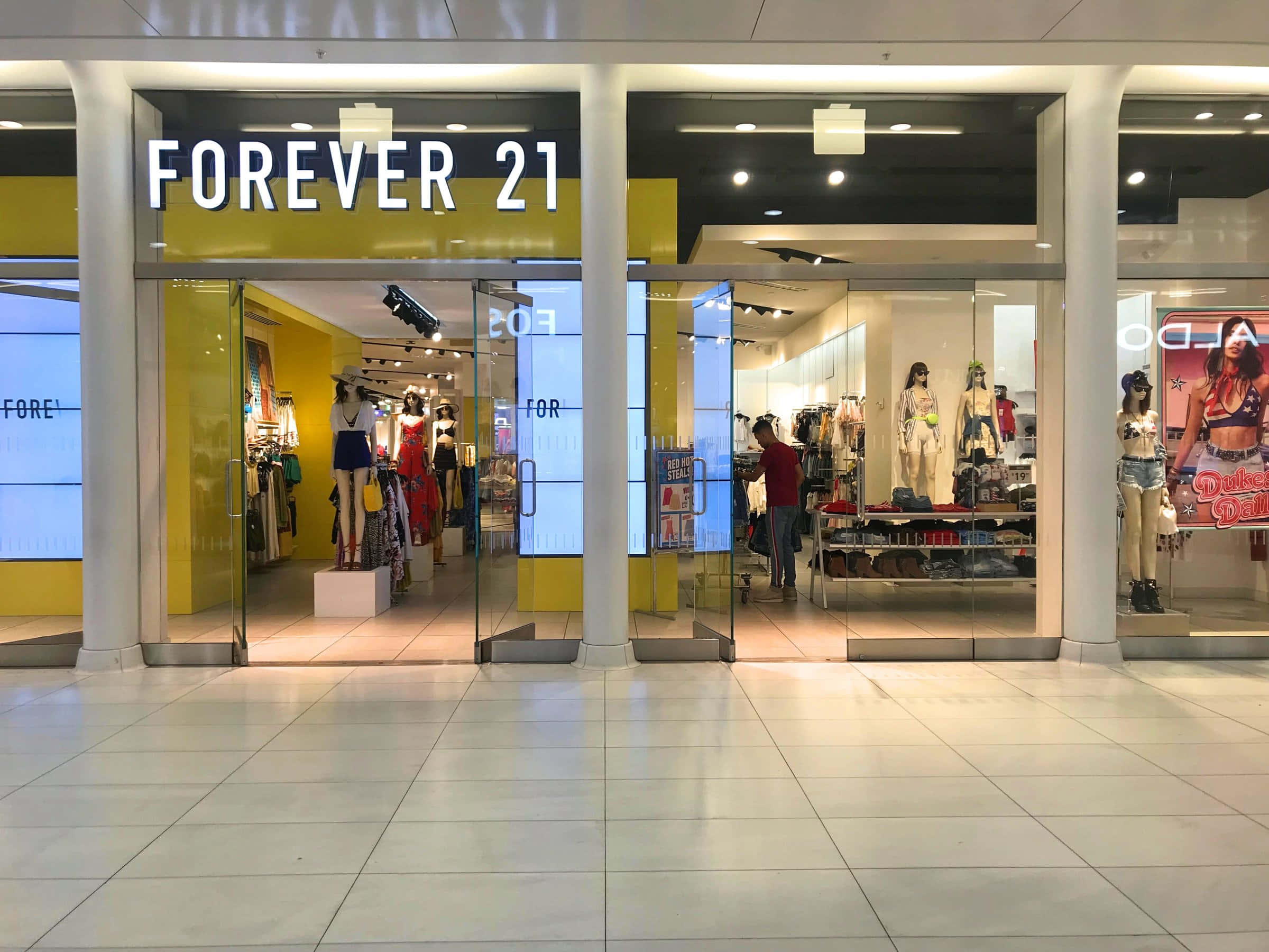 永遠21 桌布 壁紙 — 購物中心內一間Forever 21商店燈光明亮的入口，人體模型展示色彩繽紛的服裝，一名購物者在店內瀏覽。