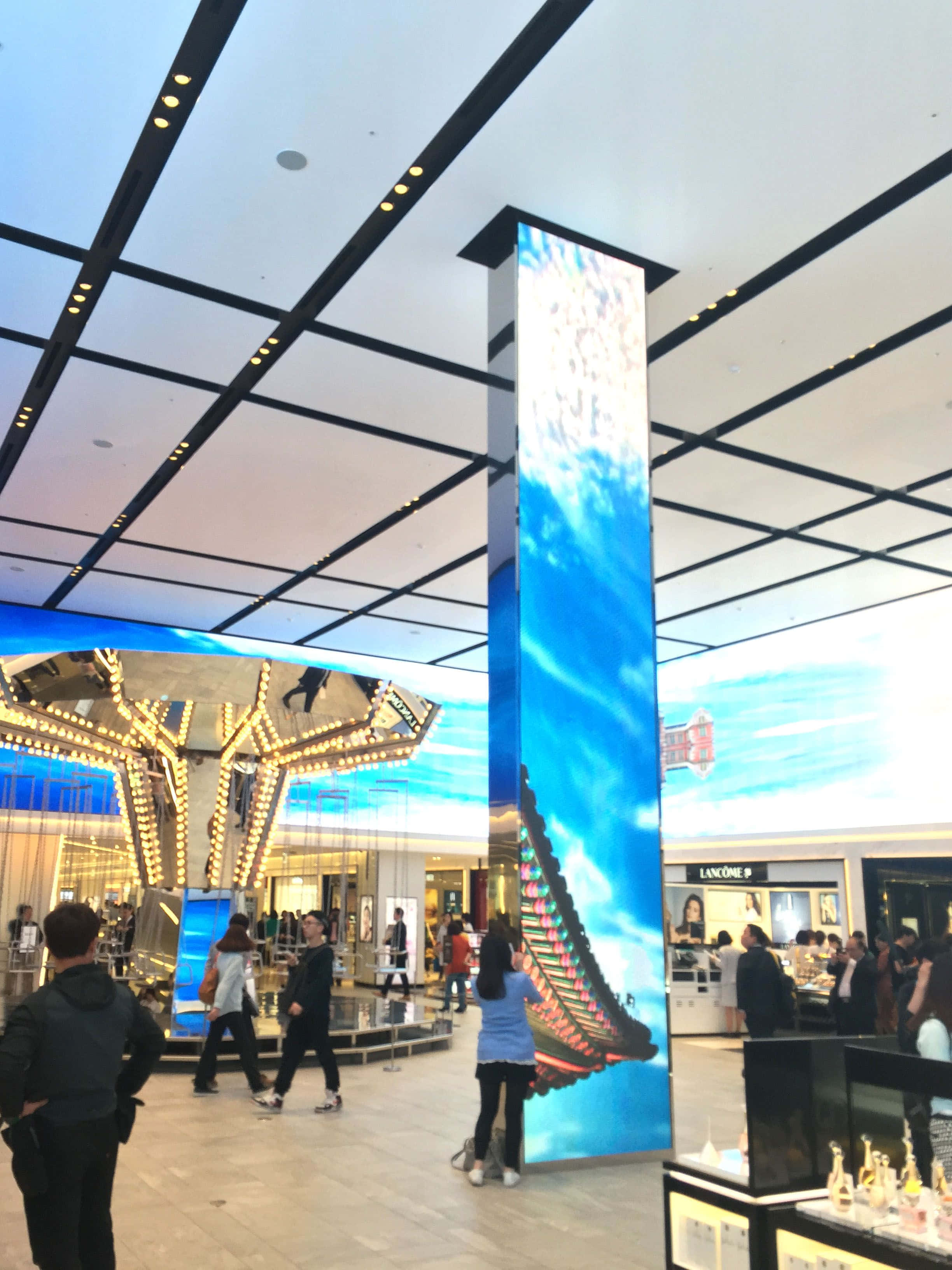Carnival Vista 桌布 壁紙 — 購物者在現代購物中心中庭，於高聳的數位螢幕下漫步，螢幕顯示著鮮豔的藍天和傳統屋頂。