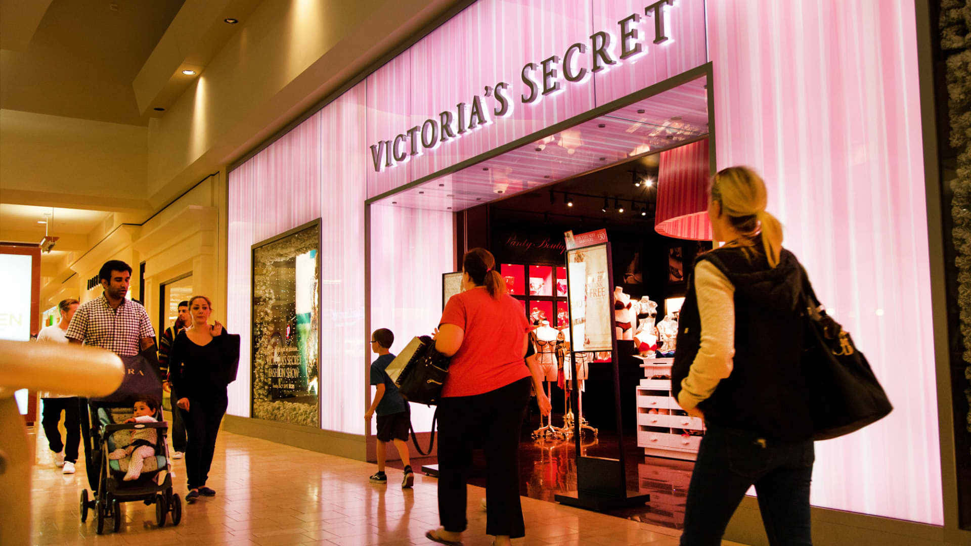 維多利亞的秘密 桌布 壁紙 — 購物者漫步經過一間燈光明亮的Victoria's Secret商店，店內有粉紅色垂直照明和展示內衣的玻璃窗。