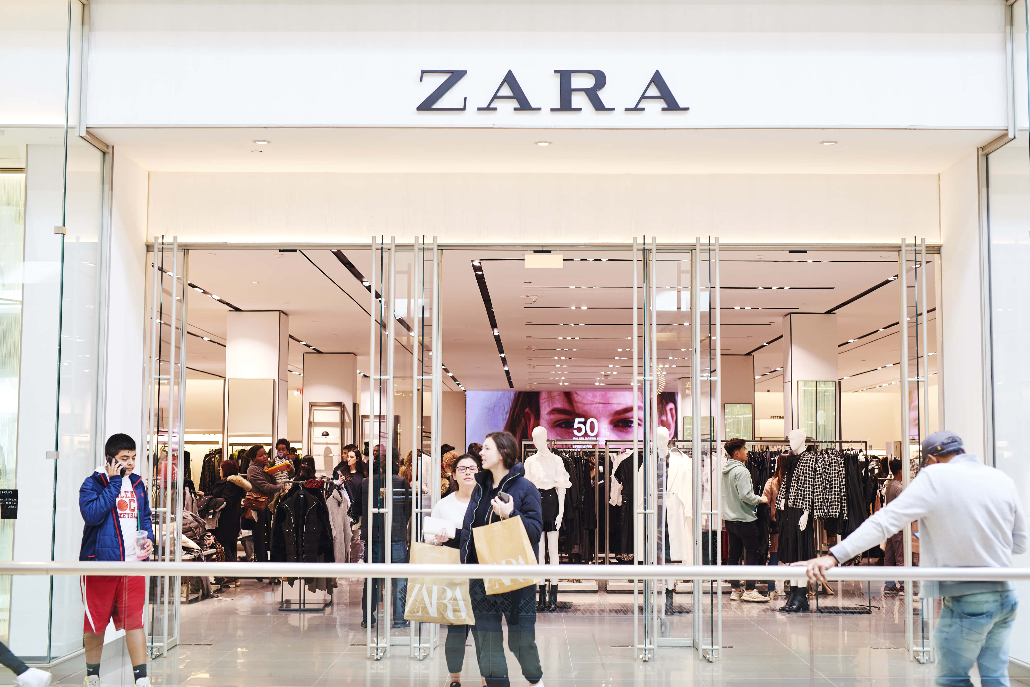 Zara 桌布 壁紙 — 購物者瀏覽並進入一間明亮現代的Zara商店，店內有玻璃門和可見的衣架。