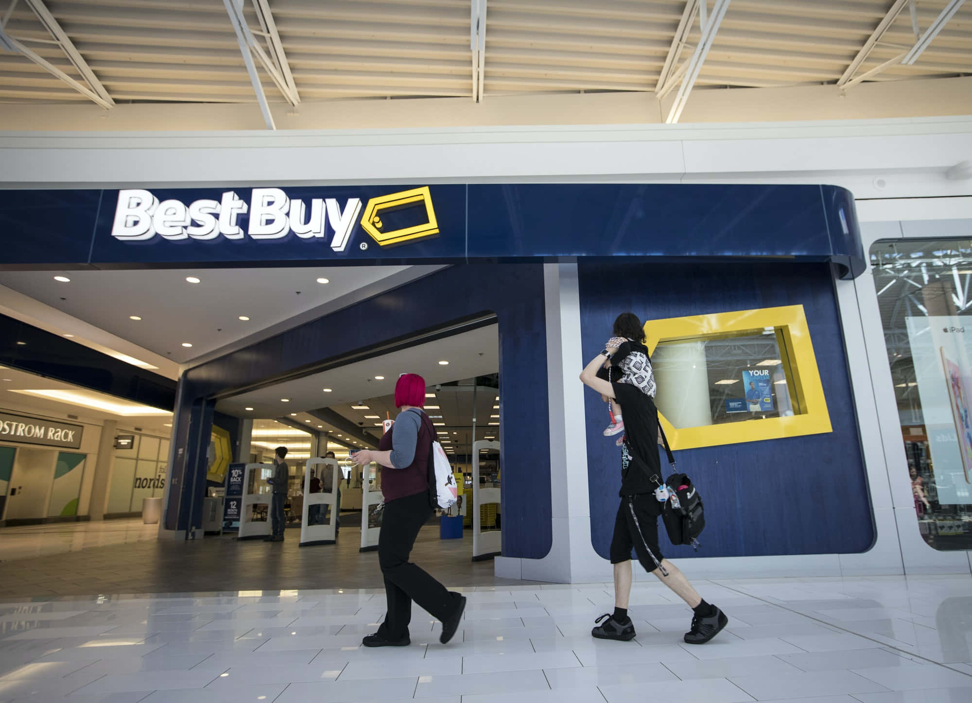 Best Buy 桌布 壁紙 — 購物者經過一間Best Buy商店入口，在一個光線充足、鋪有瓷磚地板和頂燈的購物中心內。