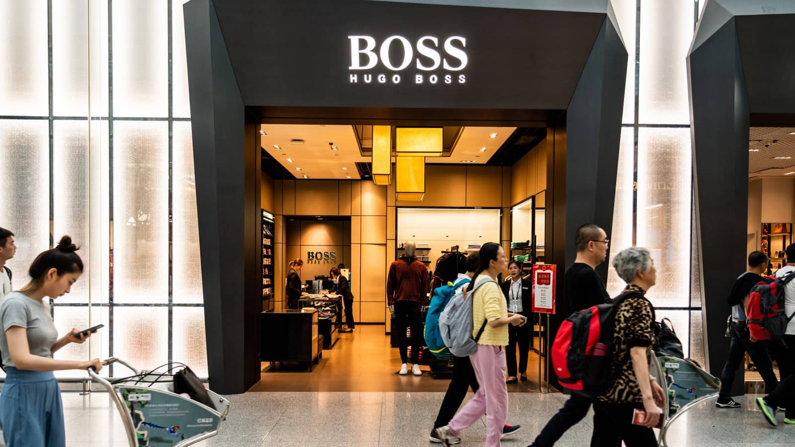 Clothes 桌布 壁紙 — 購物者經過一間現代化的Hugo Boss店鋪入口，店鋪擁有明亮的玻璃牆和溫暖的室內燈光。