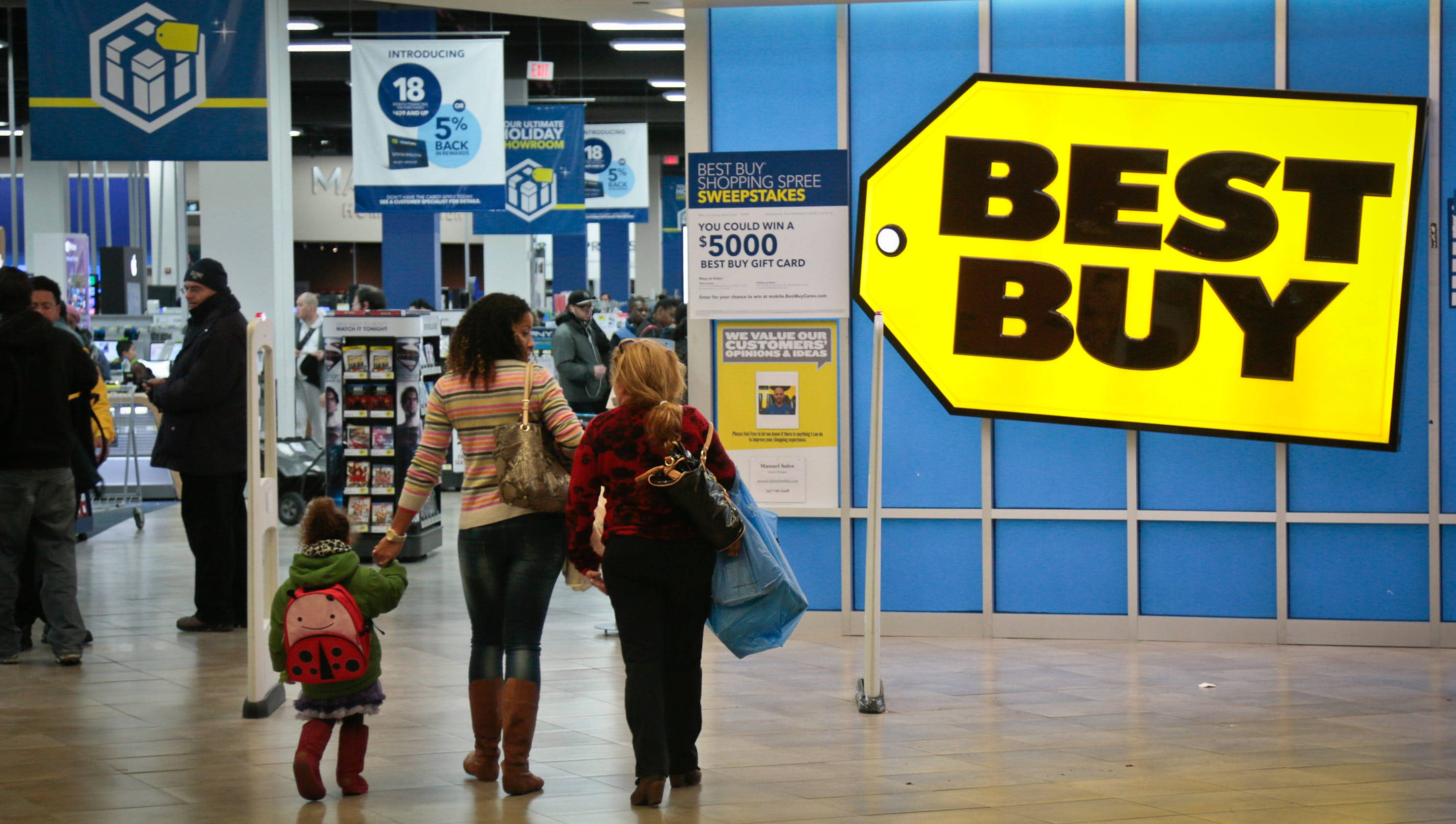 buy 桌布 壁紙 — 購物者走過光線明亮的 Best Buy 商店，頭頂上有大型黃色標籤標誌和促銷橫幅。
