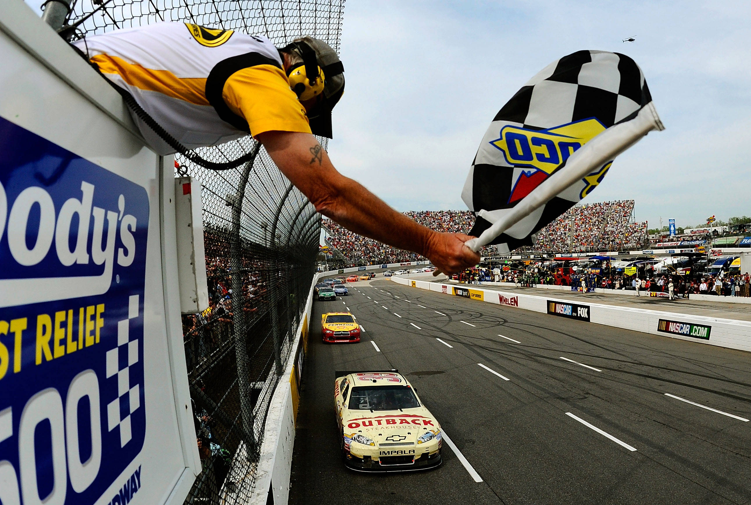 Checkered Flag 桌布 壁紙 — 賽事工作人員揮動方格旗，一輛NASCAR賽車在晴朗天空下衝過終點線。