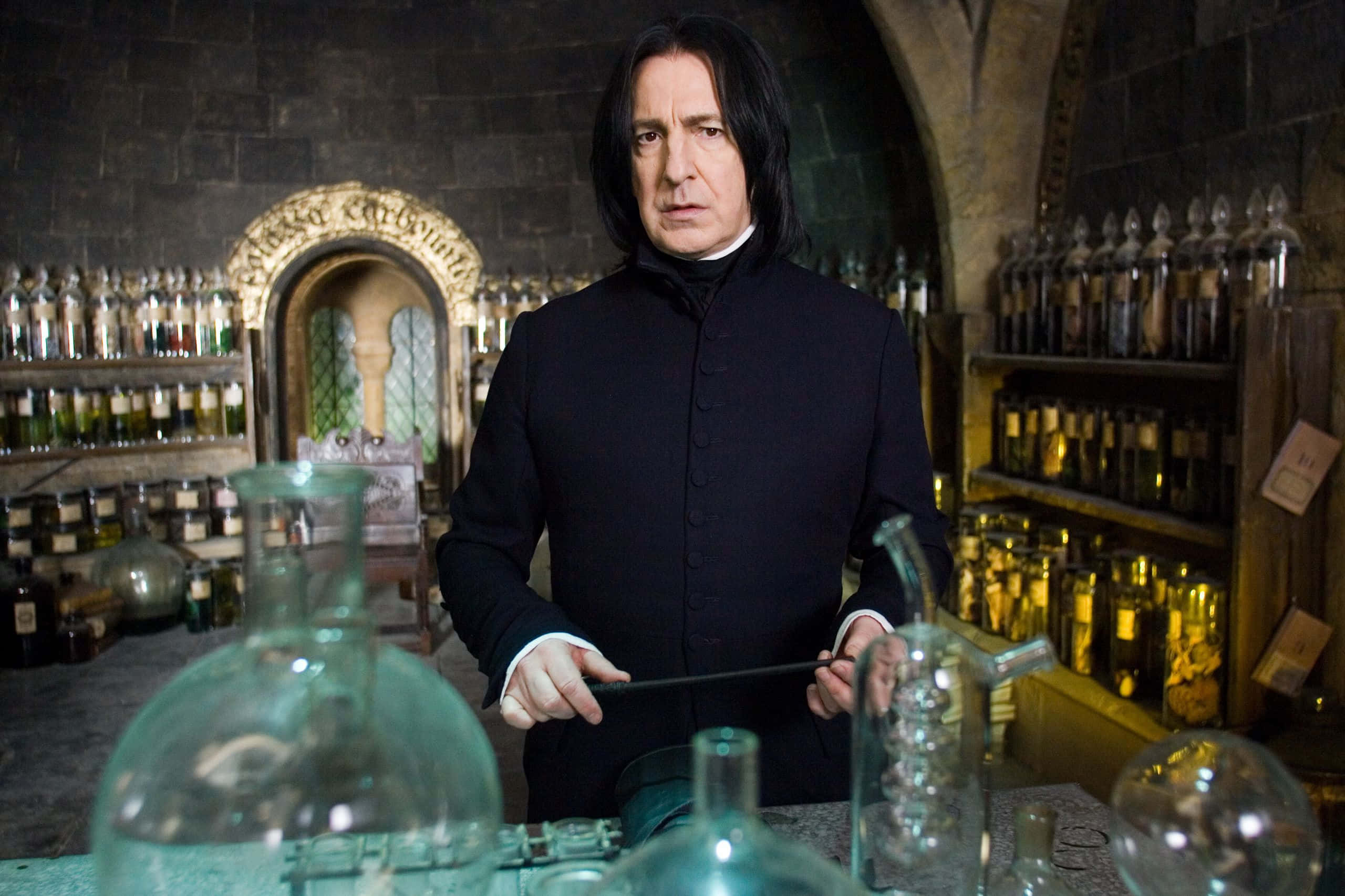 The Hogwarts Potions Class 桌布 壁紙 — 賽佛勒斯·石內卜站在燈光昏暗的魔藥學教室，手持魔杖，周圍是玻璃瓶和裝滿魔法原料的架子。