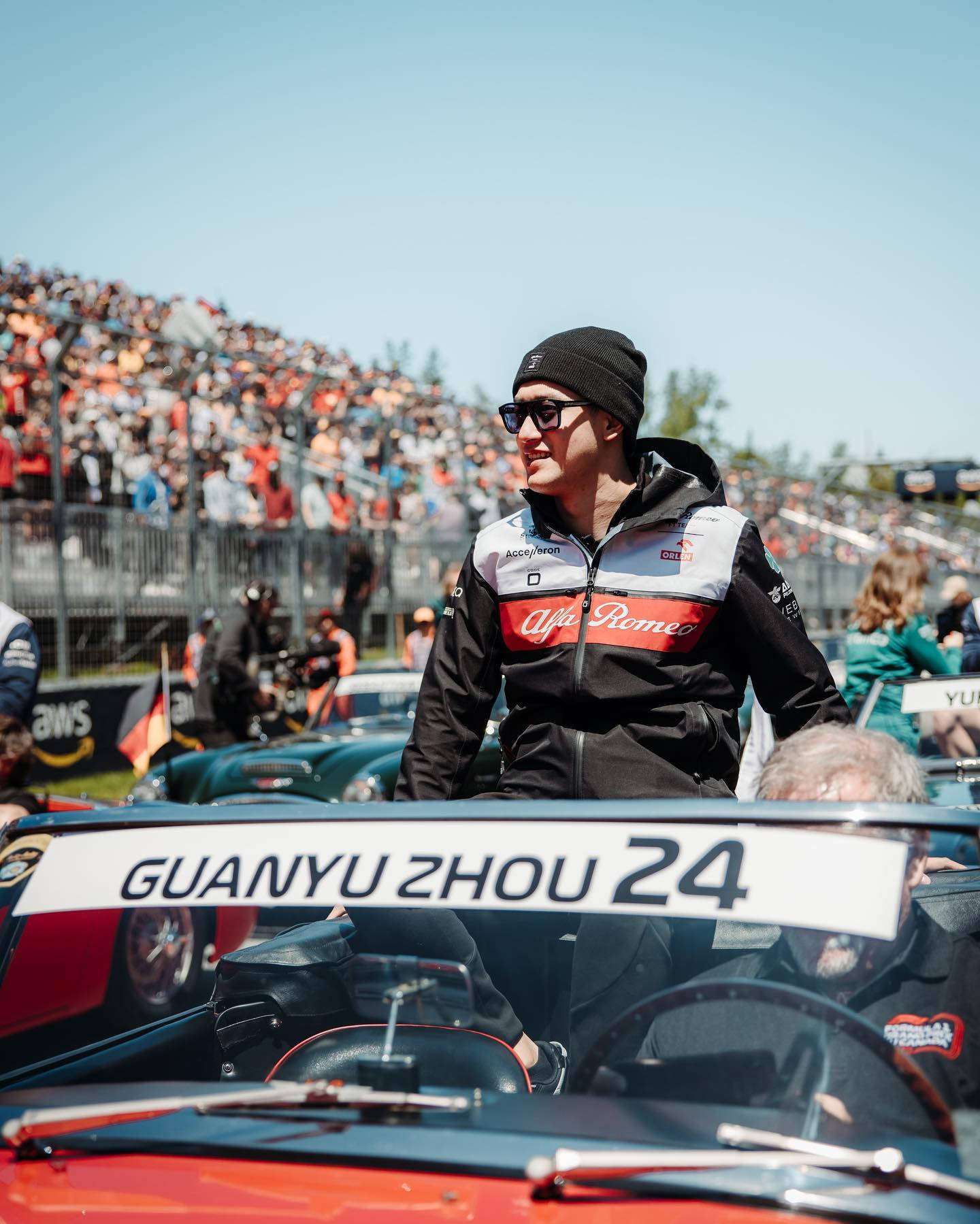 F1 桌布 壁紙 — 賽車手周冠宇在他的愛快羅密歐駕駛艙中，在陽光明媚的賽道上，看台上的粉絲歡呼著。