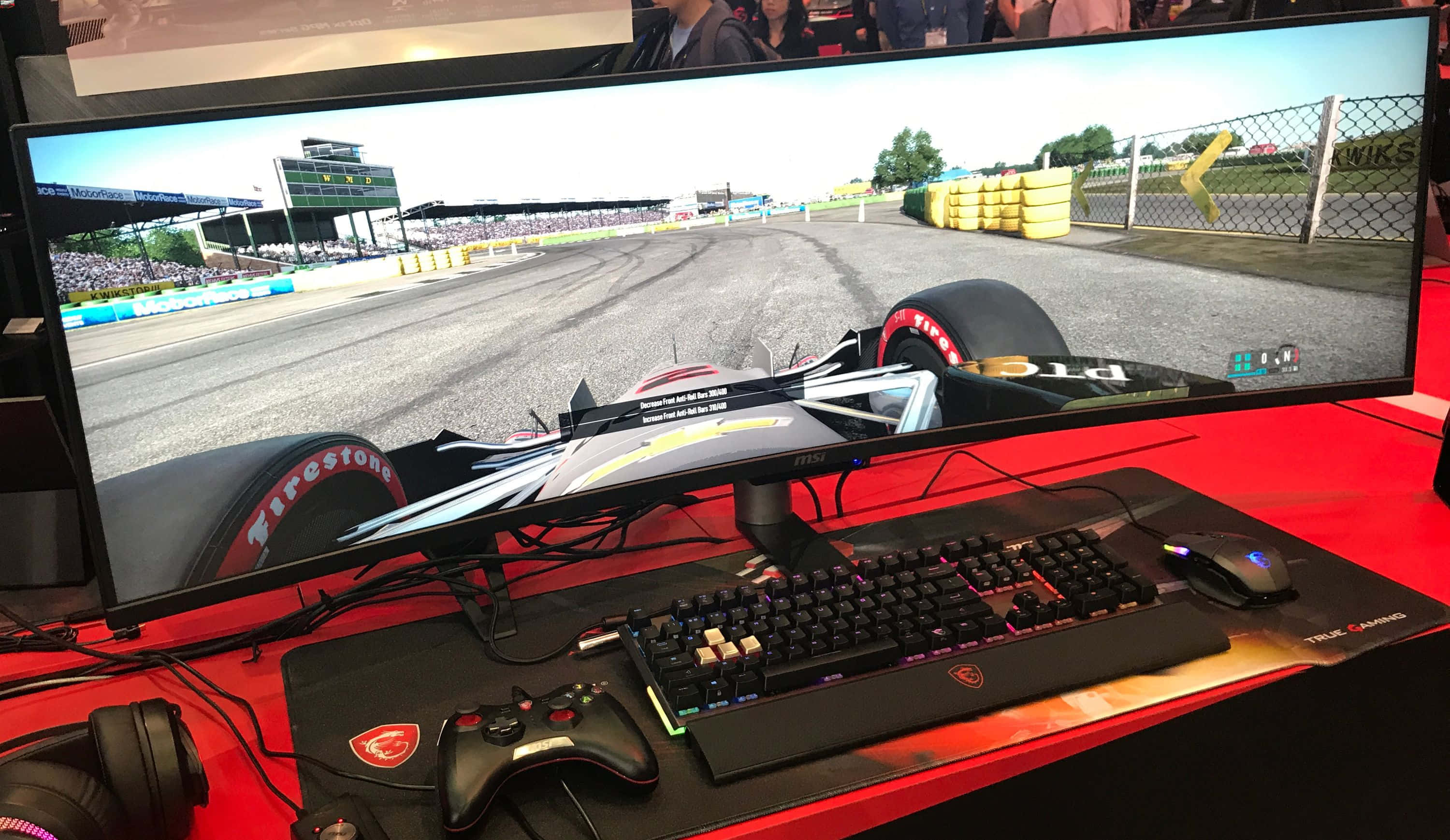 msi 2560x1080 桌布 壁紙 — 賽車遊戲顯示在曲面顯示器上，遊戲設置包括鍵盤、滑鼠和控制器，放在紅色桌子上。