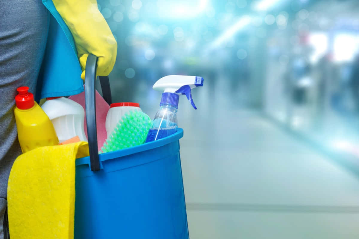 Cleaning 桌布 壁紙 — 走廊中一個人手持的藍色清潔桶特寫，桶內裝有噴霧瓶、海綿和一條黃色抹布。