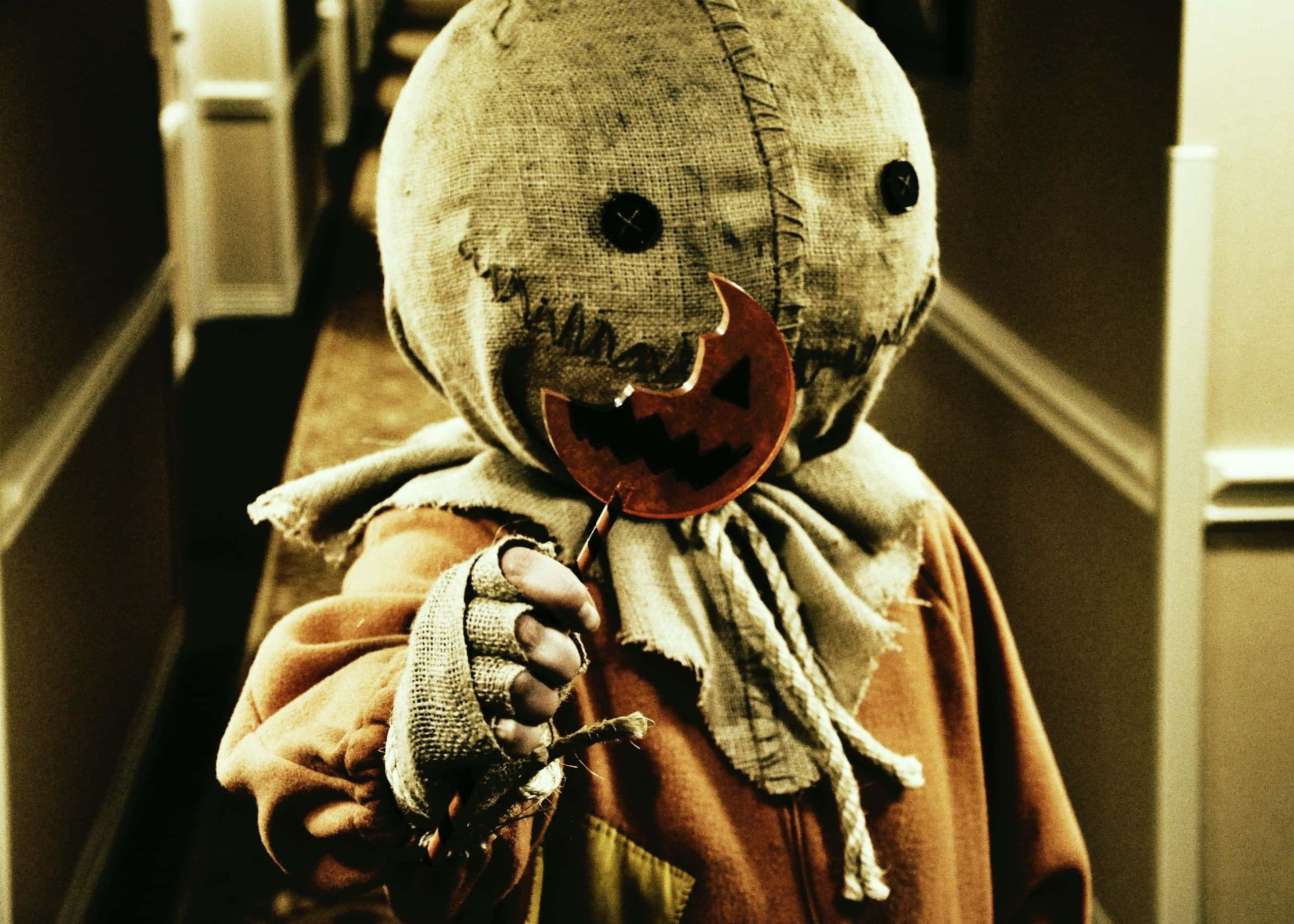 Monster Movies 桌布 壁紙 — 走廊裡一個縫製的稻草人小孩拿著一個南瓜棒棒糖，露出猙獰的笑容。