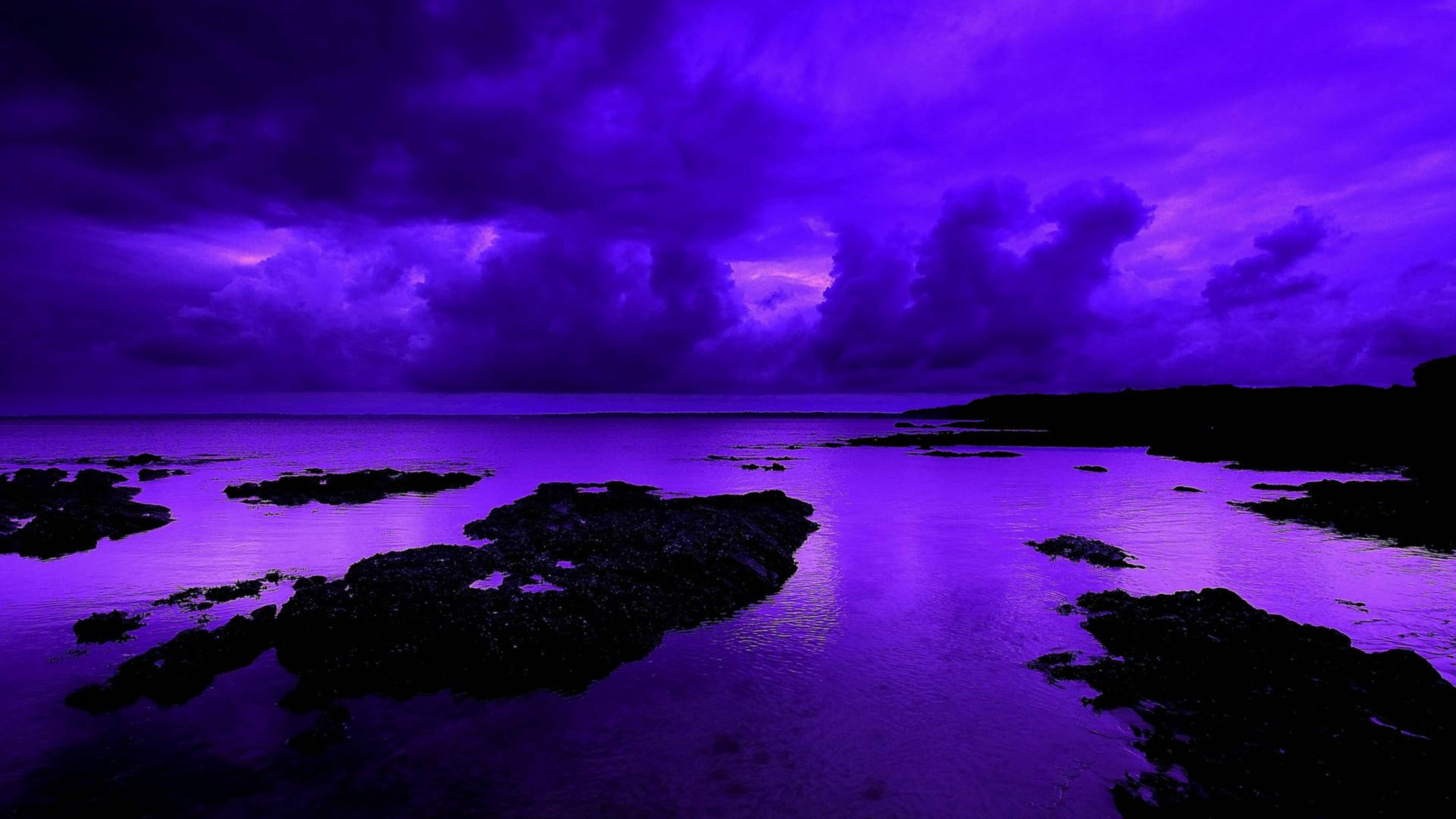 Violet Aesthetic 桌布 壁紙 — 超現實的紫色調海岸場景，烏雲在平靜的水面上倒映，襯托出岩石的剪影。