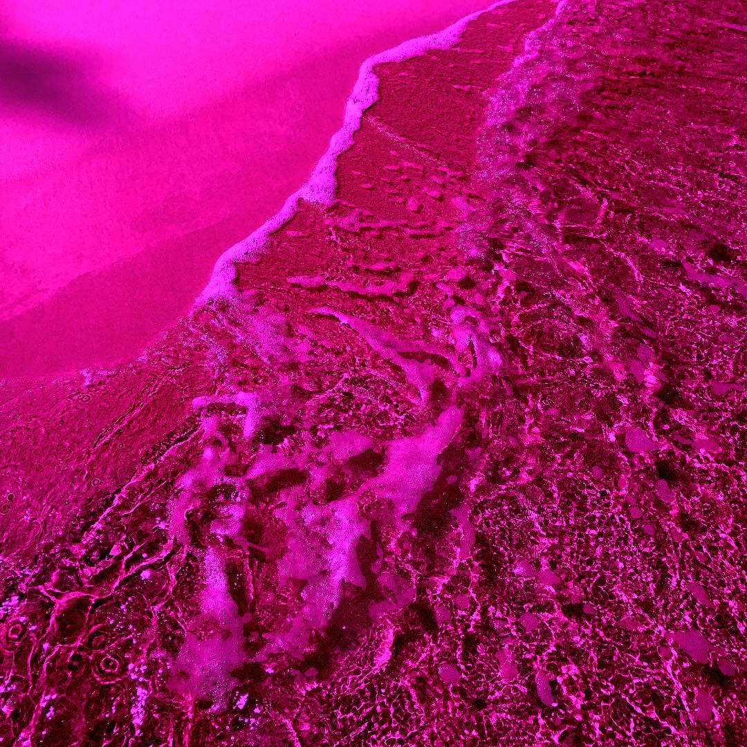 Hot Pink Aesthetic 桌布 壁紙 — 超現實的航拍視角，海浪拍打著被濃烈洋紅和紫紅色調浸染的海灘。