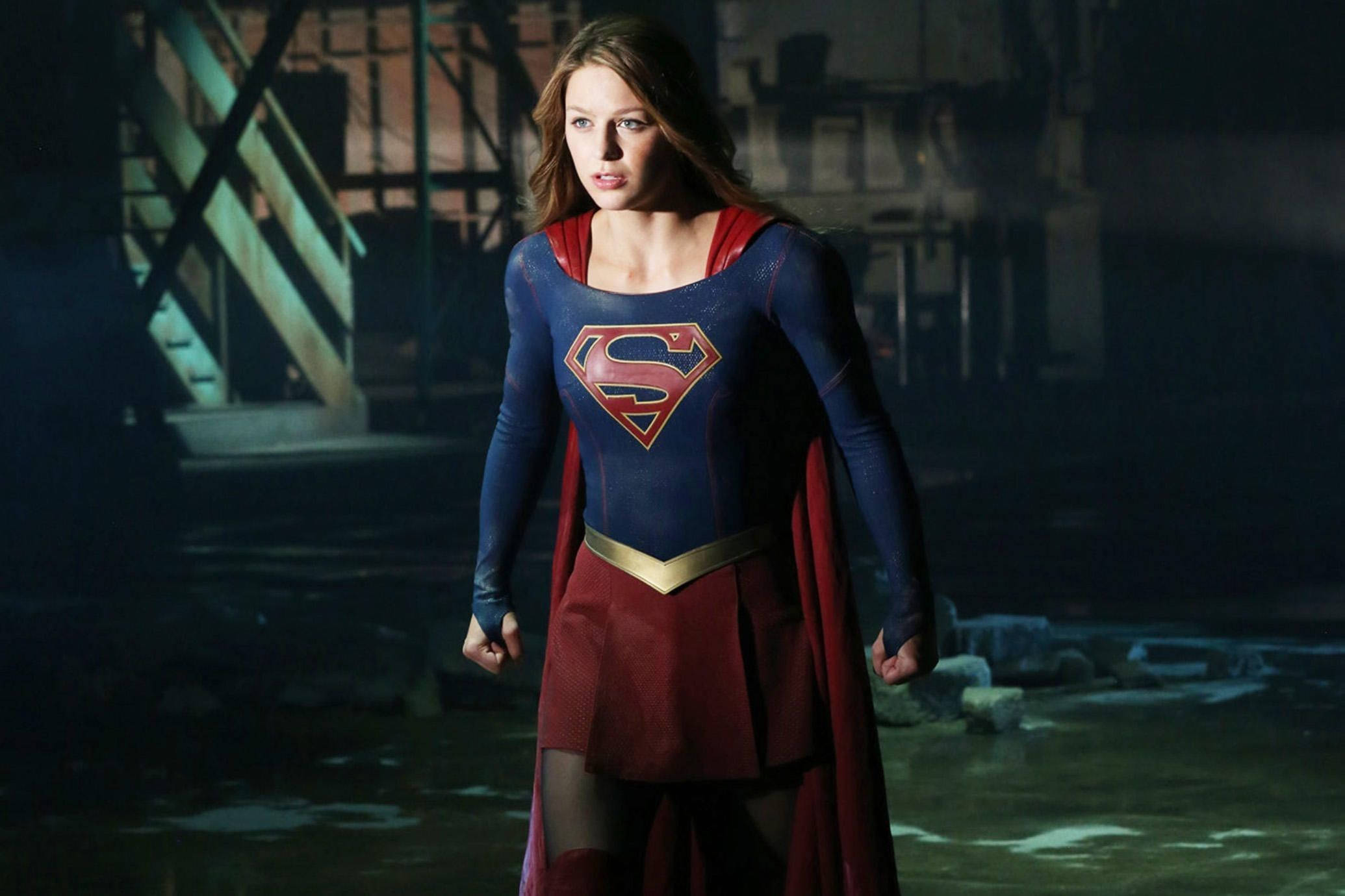 Superwoman 桌布 壁紙 — 超級女孩在黑暗的工業廢墟中擺出姿勢，身穿標誌性的藍色和紅色服裝，眼神堅定。