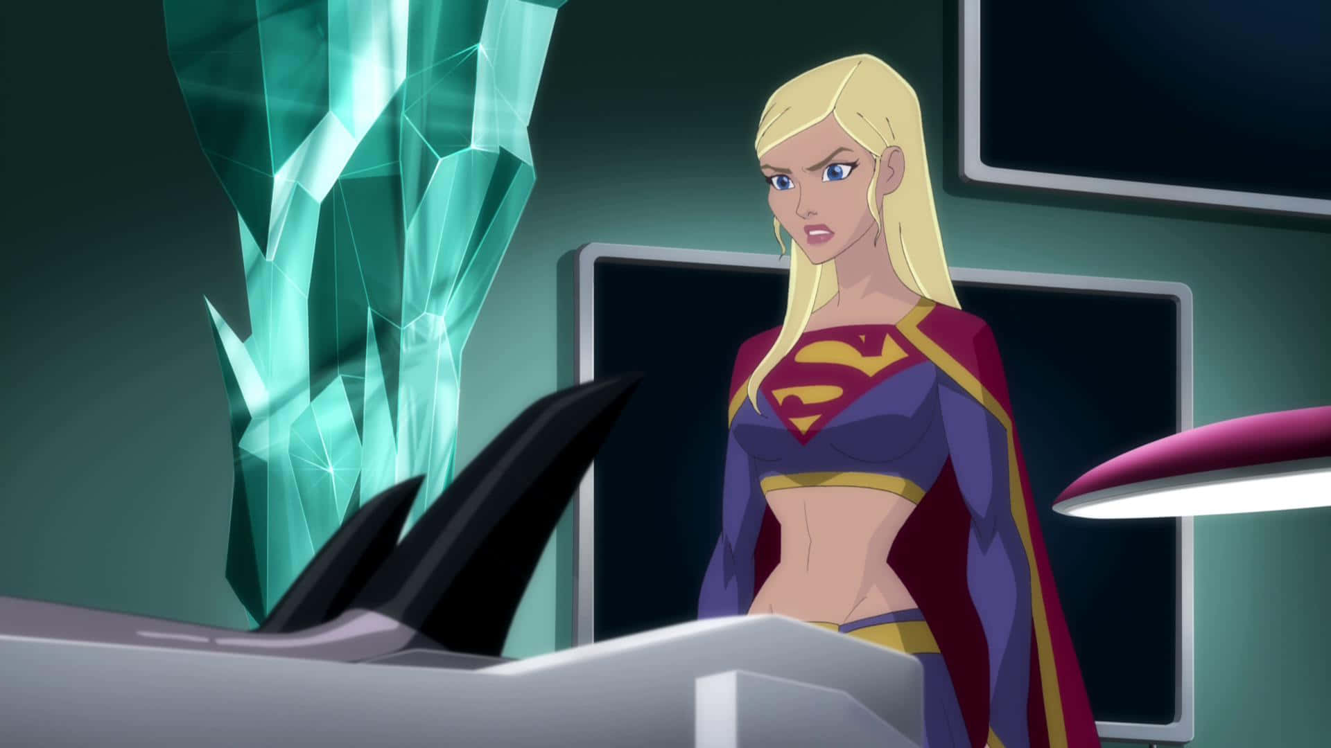 Superman Unbound 桌布 壁紙 — 超級女孩站在一個高科技實驗室裡，旁邊有一塊發光的綠色水晶，周圍有顯示器和未來主義設備。