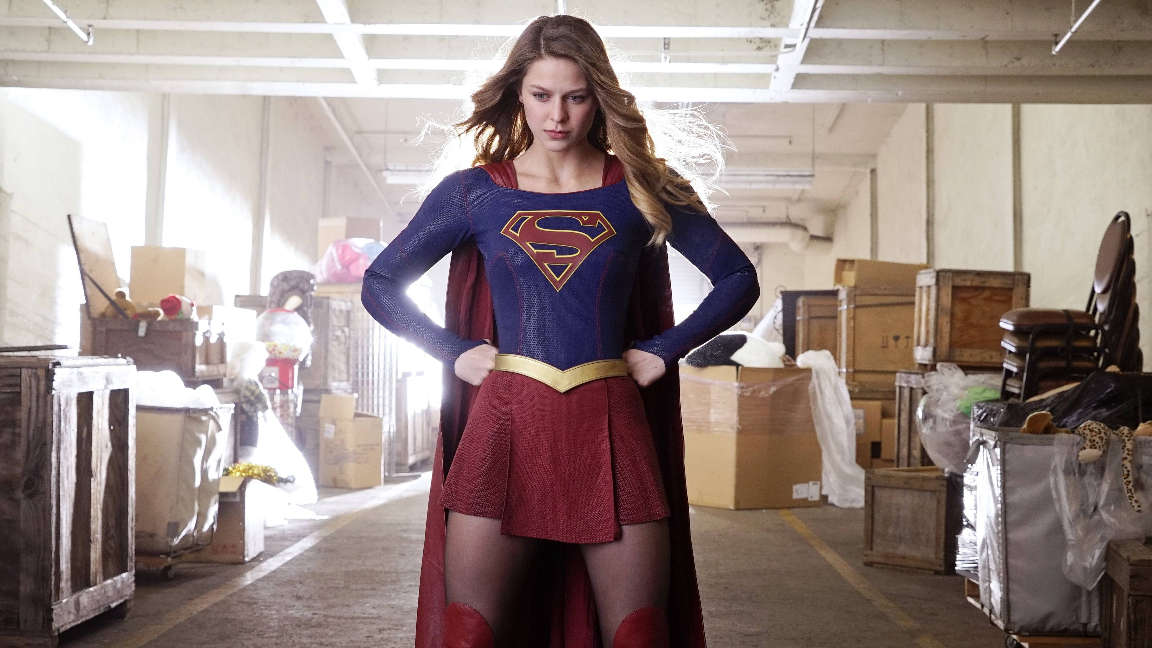 Superwoman 桌布 壁紙 — 超級女孩自信地站在一個雜亂的倉庫中，穿著她標誌性的藍色和紅色服裝，表情堅定。