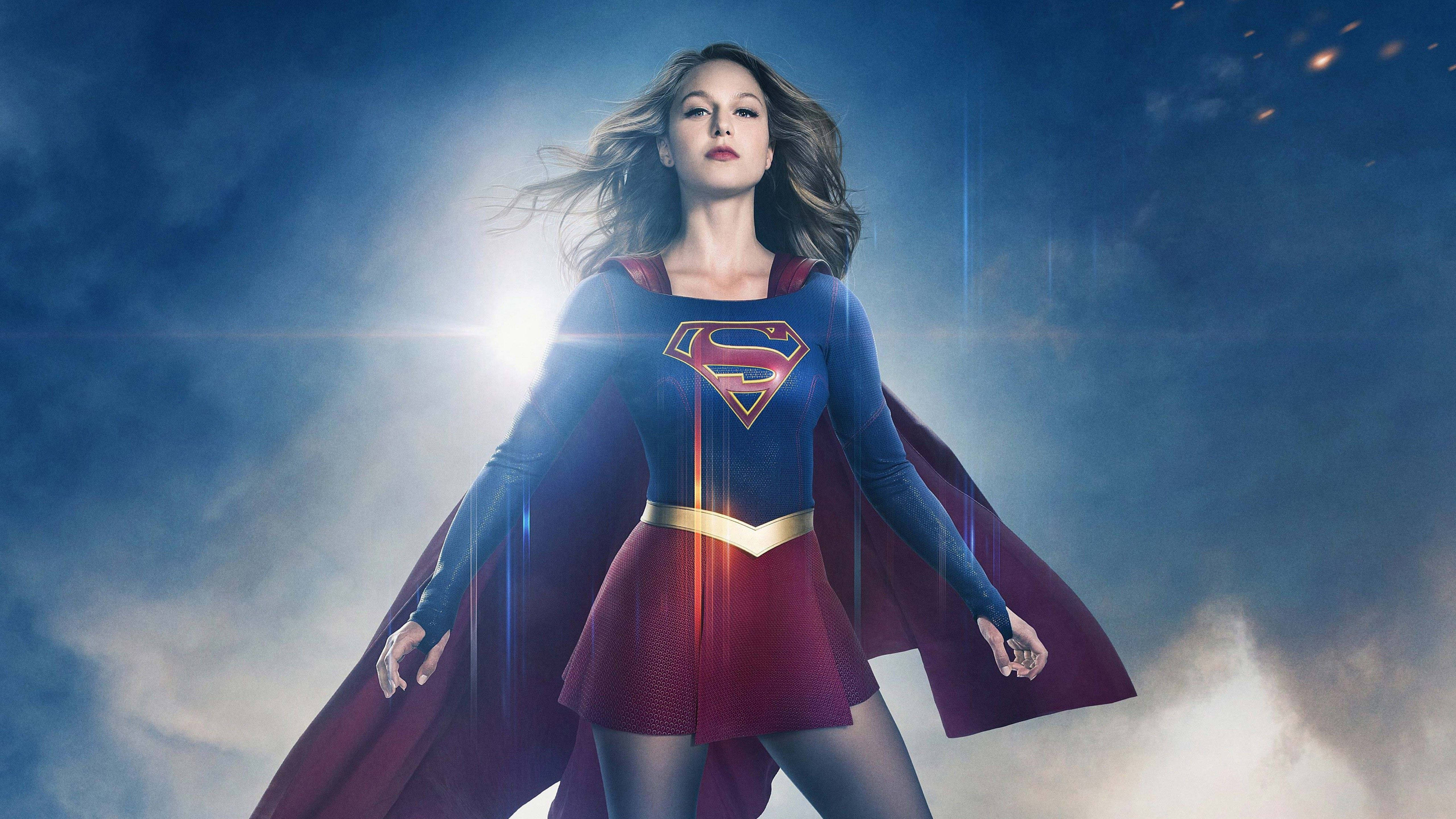 Superwoman 桌布 壁紙 — 超級女英雄以英雄姿態站在戲劇性的天空下，陽光從她飄逸的斗篷後方迸射而出。