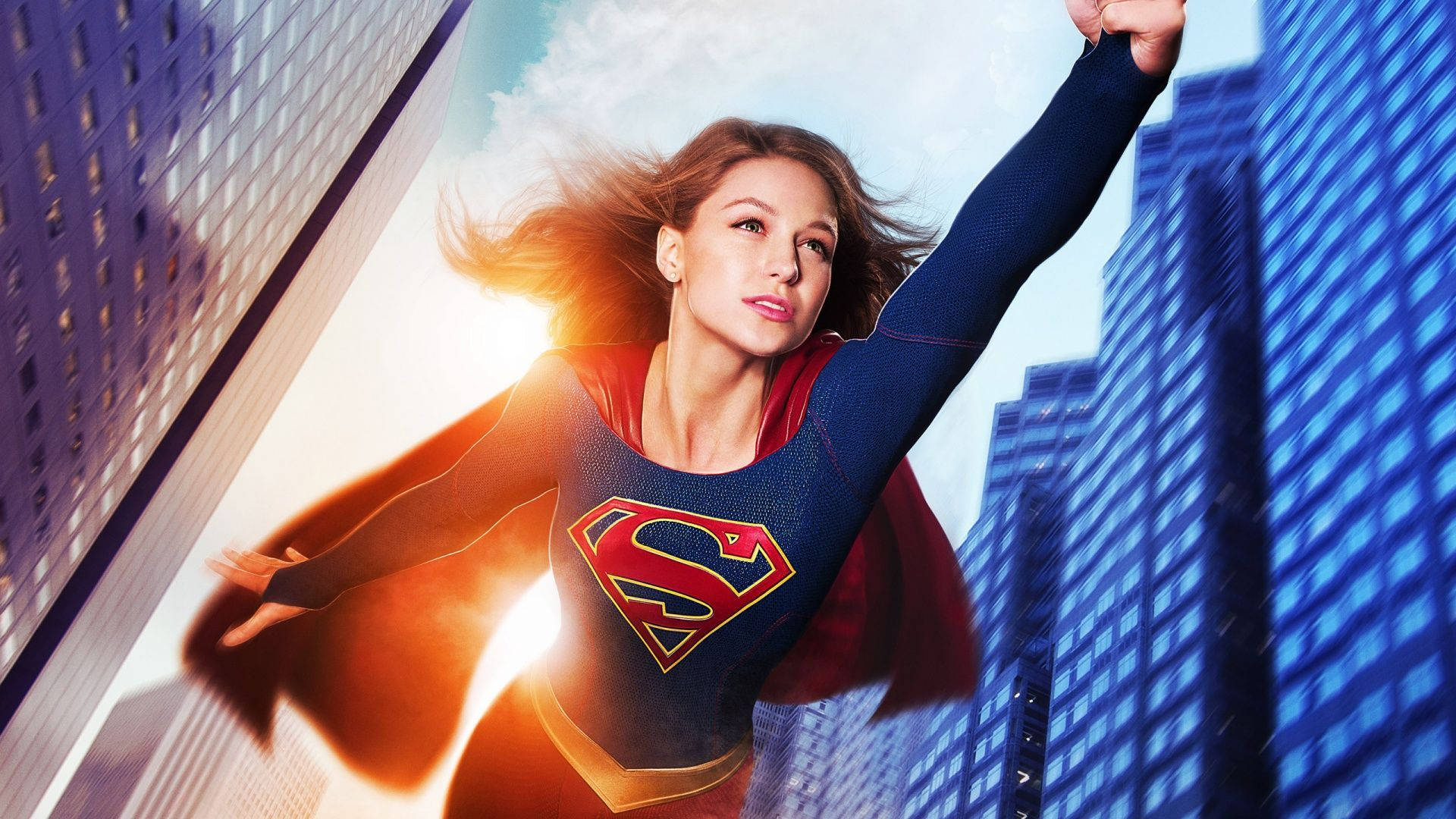 Superwoman 桌布 壁紙 — 超級女超人翱翔於高聳的摩天大樓之間，風吹拂她的頭髮，陽光在她身後綻放。