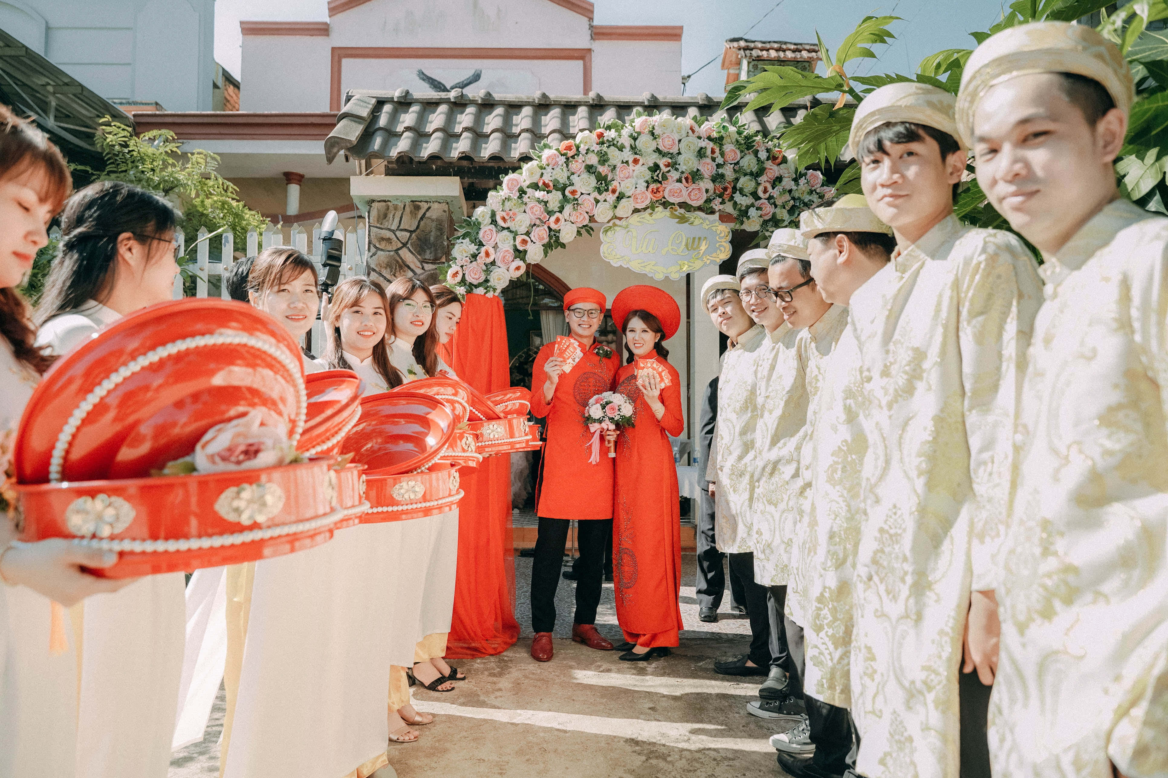 Groomsman 桌布 壁紙 — 越南婚禮遊行中，新娘身穿紅色奧黛，新郎身穿白色奧黛，在花拱下，伴隨者手持紅色儀式托盤。