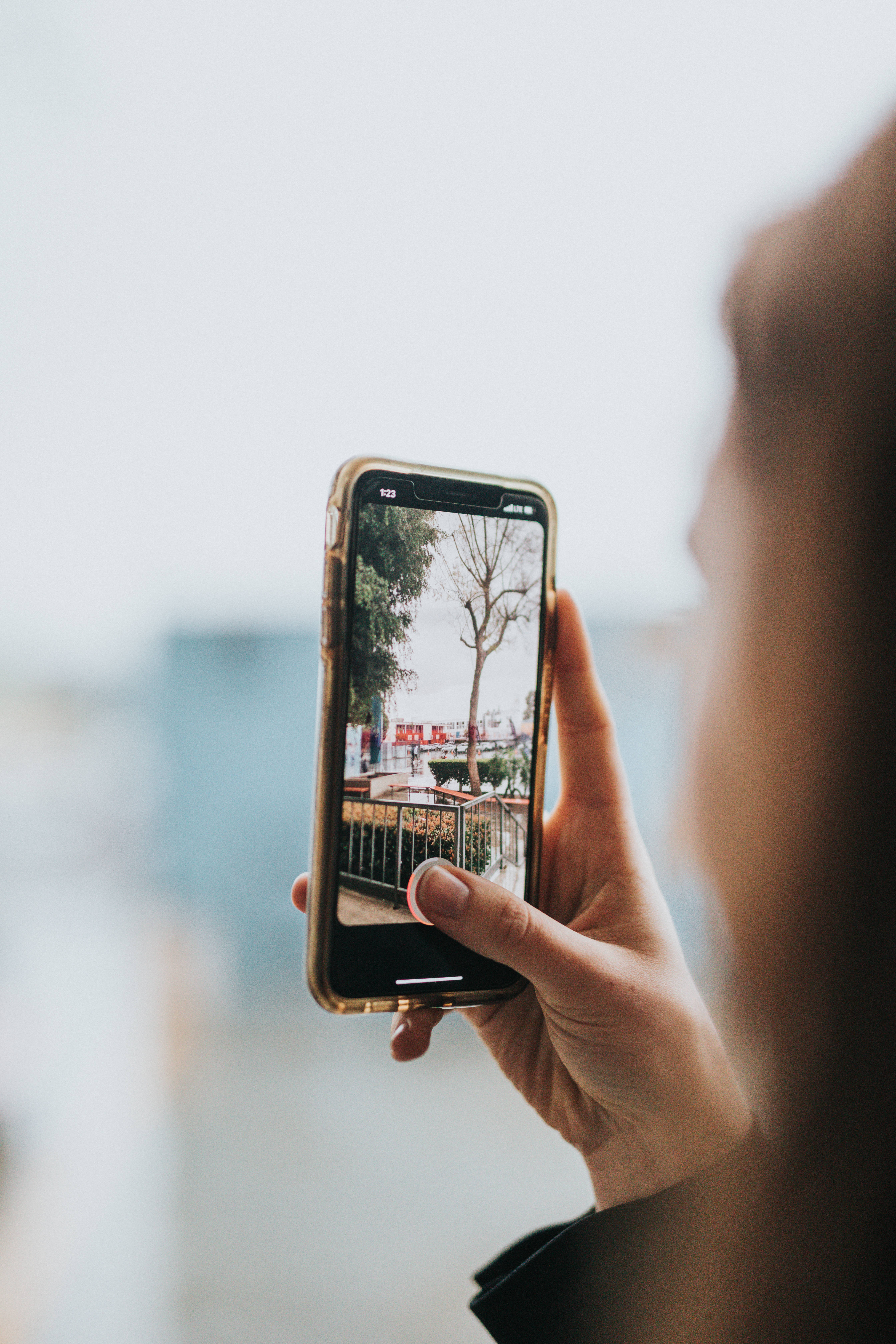 iPhone 攝影 桌布 壁紙 — 越過肩膀的視角，一隻手拿著智慧型手機，顯示一張有樹木和紅色標誌的陽台照片。