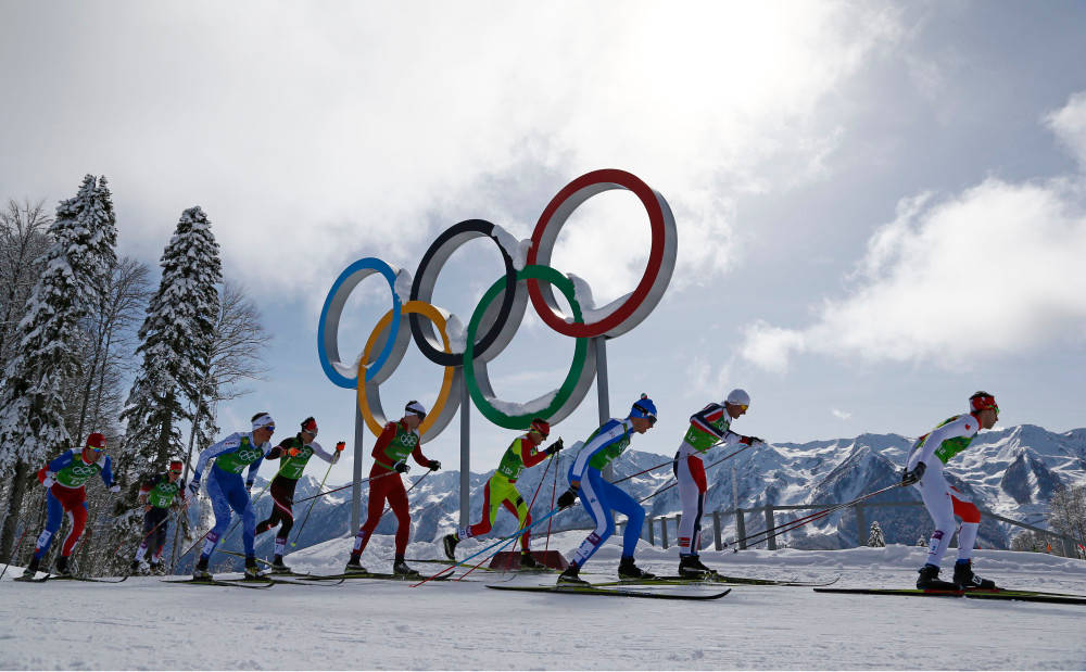 Winter Olympics 桌布 壁紙 — 越野滑雪選手在陰天、白雪覆蓋的山脈和樹木背景下，競速經過巨大的奧運五環。