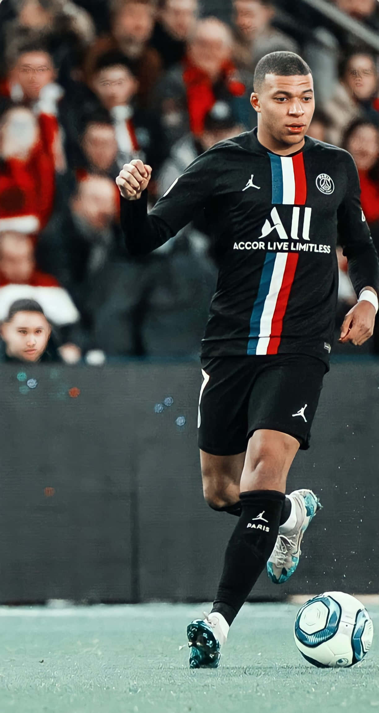 Mbappe 桌布 壁紙 — 足球員基利安·姆巴佩在綠色球場上盤球，背景是模糊的體育場觀眾。