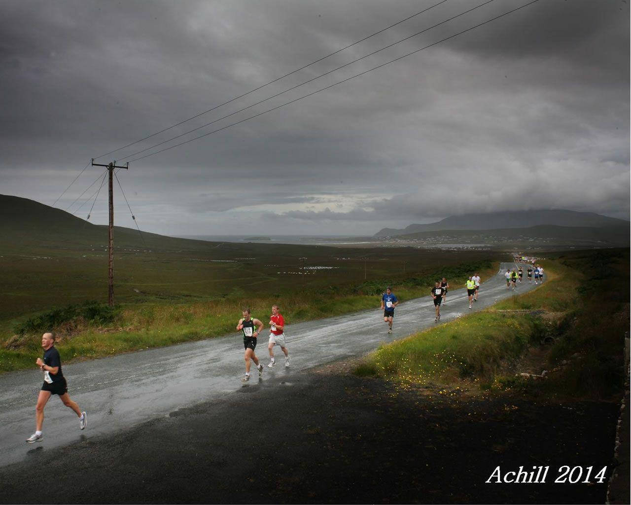 Marathon 桌布 壁紙 — 跑者在沉重的灰色雲層下沿著濕漉漉的鄉村道路奔跑，背景是遠山和海岸線。