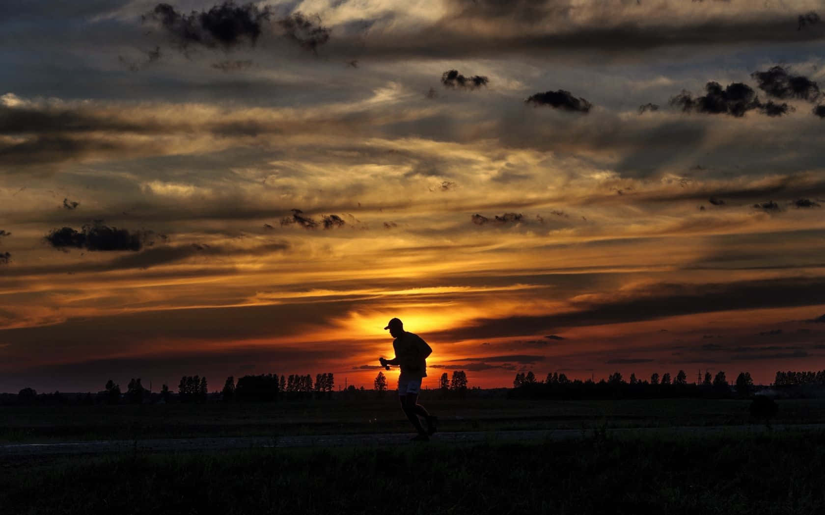 日落 桌布 壁紙 — 跑者的剪影在火紅的夕陽天空下掠過田野，天空佈滿橙色和深色雲彩。