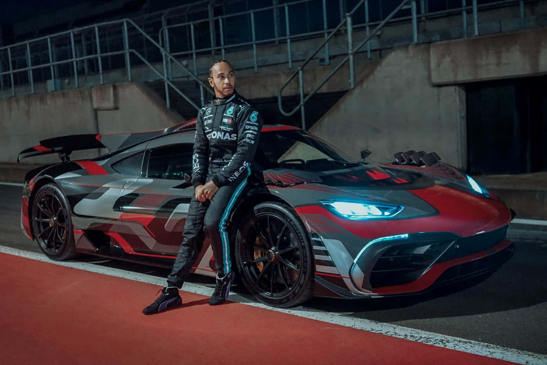 Mercedes 桌布 壁紙 — 路易斯·漢米爾頓在夜間賽道上自信地靠在一輛時尚的紅黑色AMG One超跑旁。