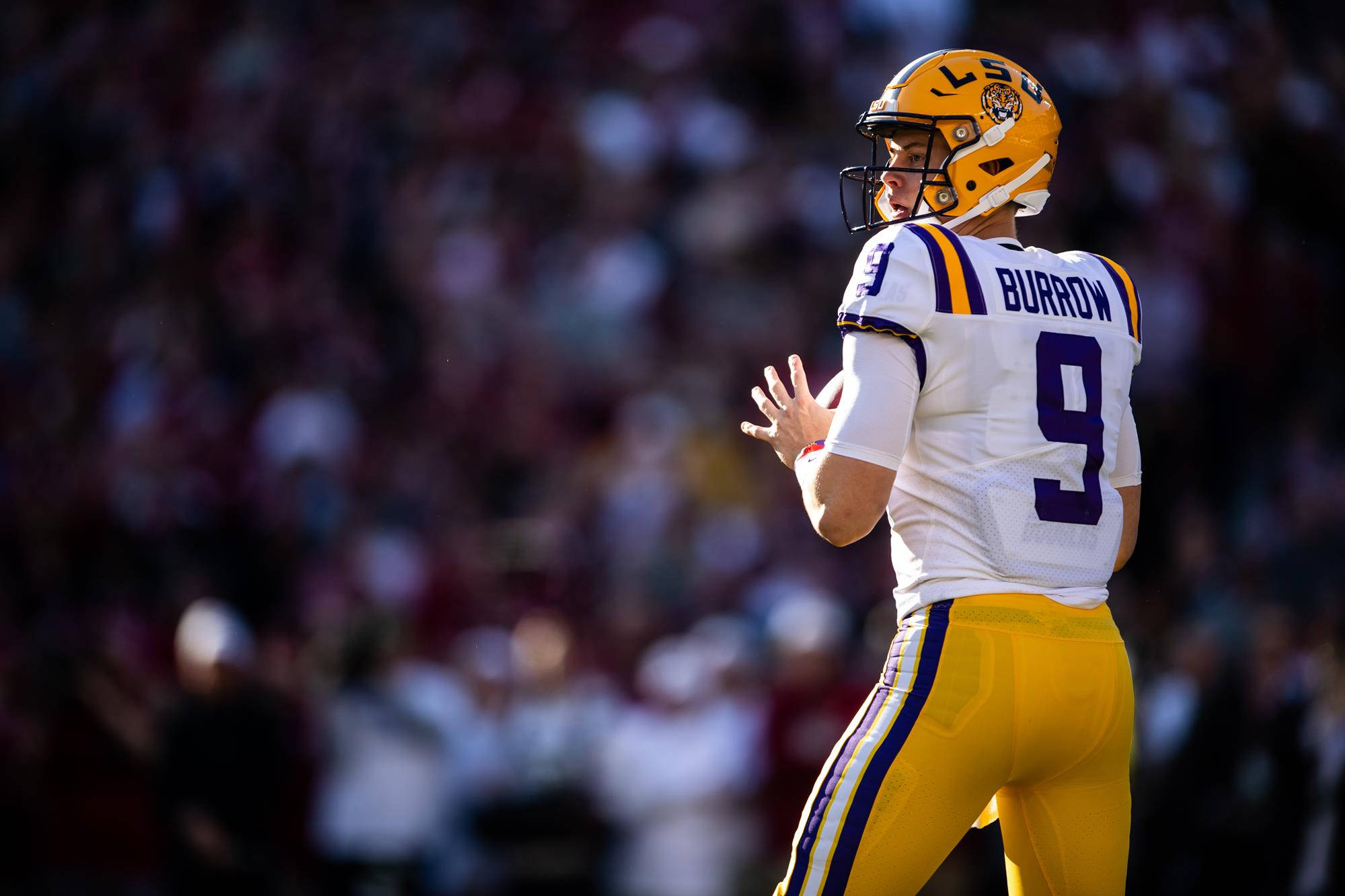 Joe Burrow 桌布 壁紙 — 身穿LSU球衣的四分衛喬·伯羅在明亮的日間足球比賽中掃視球場，背景是模糊的人群。
