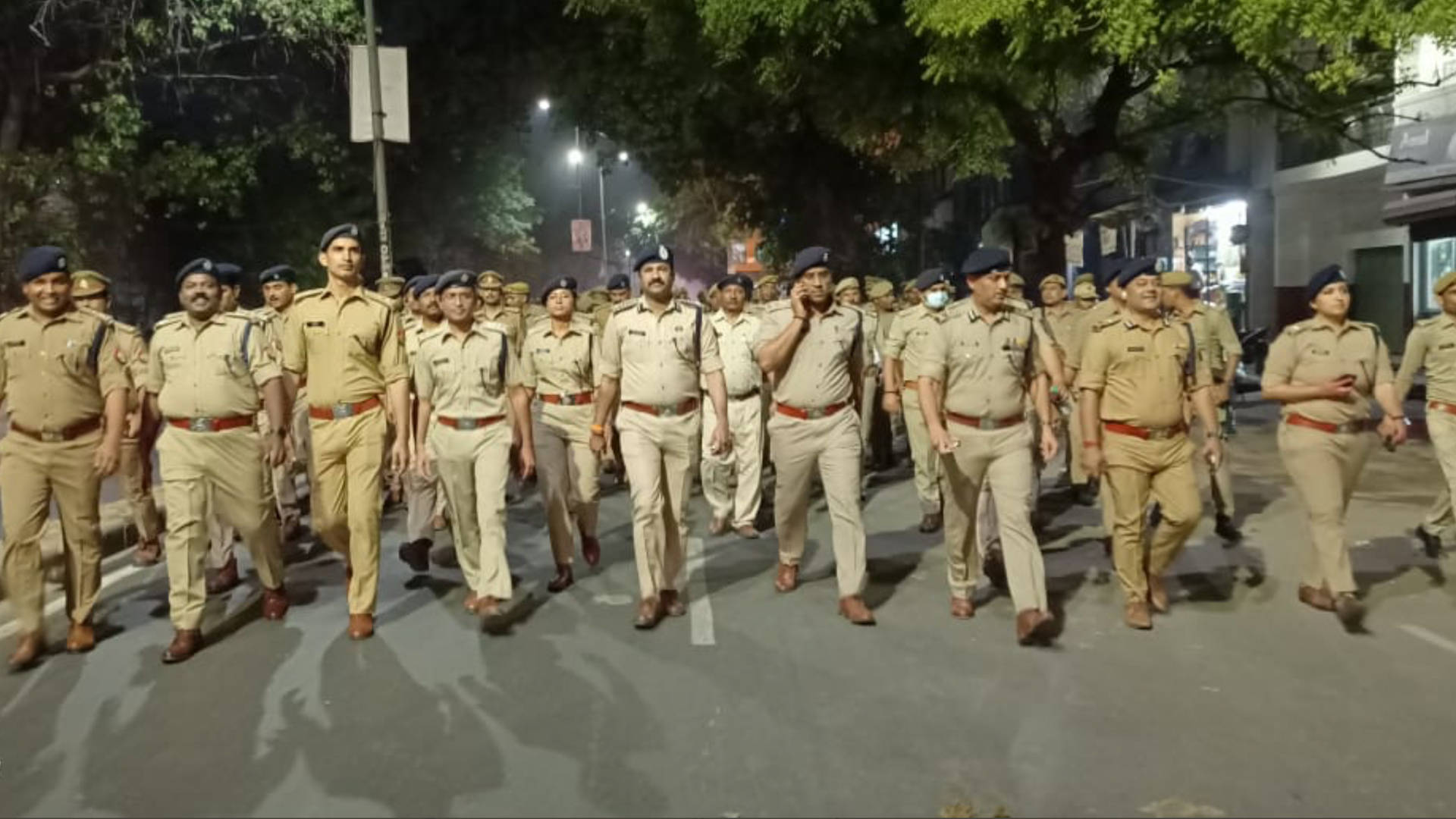 Indian 桌布 壁紙 — 身穿制服的警察在夜間路燈下，沿著綠樹成蔭的城市街道列隊行進。