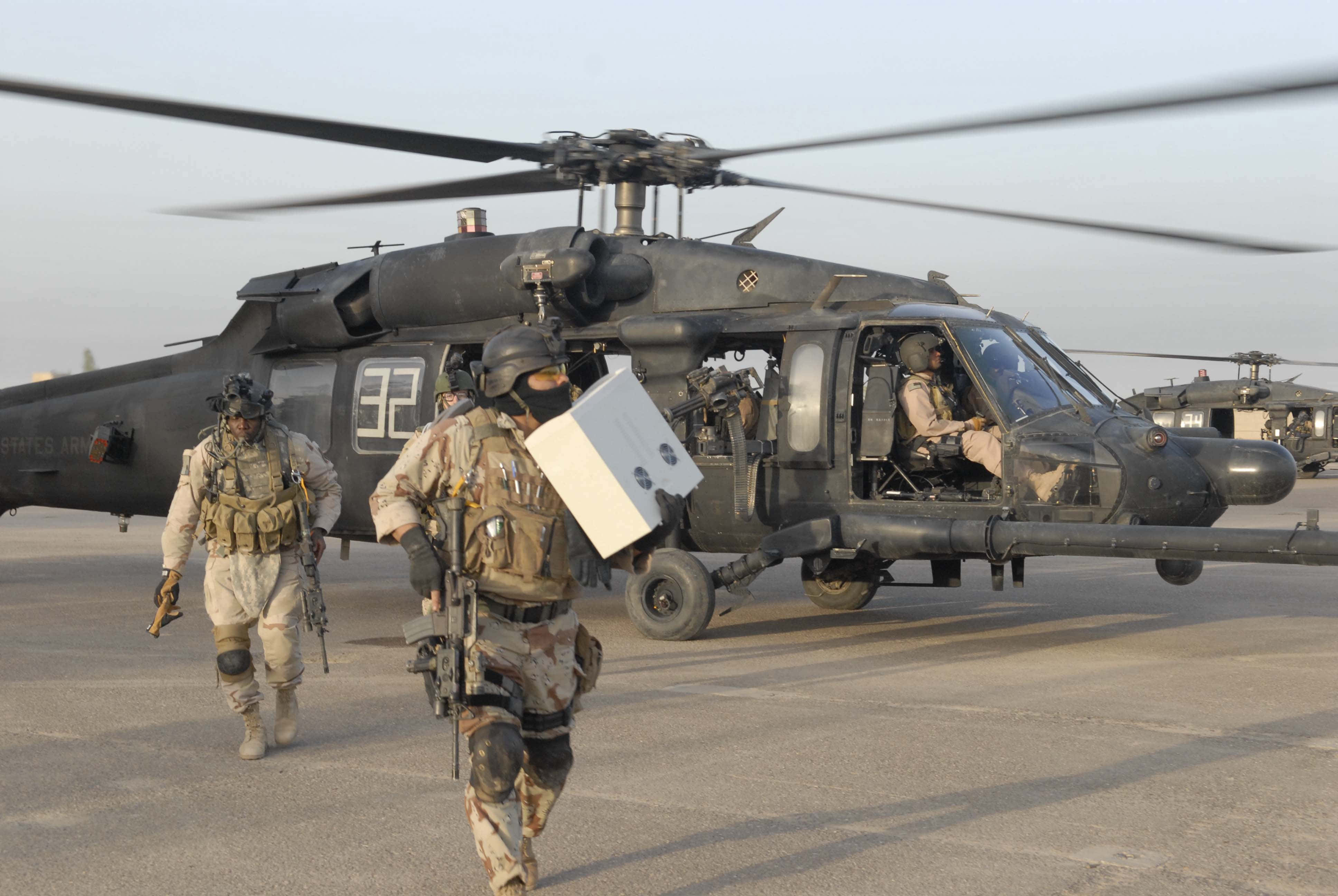 4K Helicopters 桌布 壁紙 — 身穿戰鬥裝備的士兵在塵土飛揚的機場上，向一架懸停的黑色軍用直升機運送補給品。