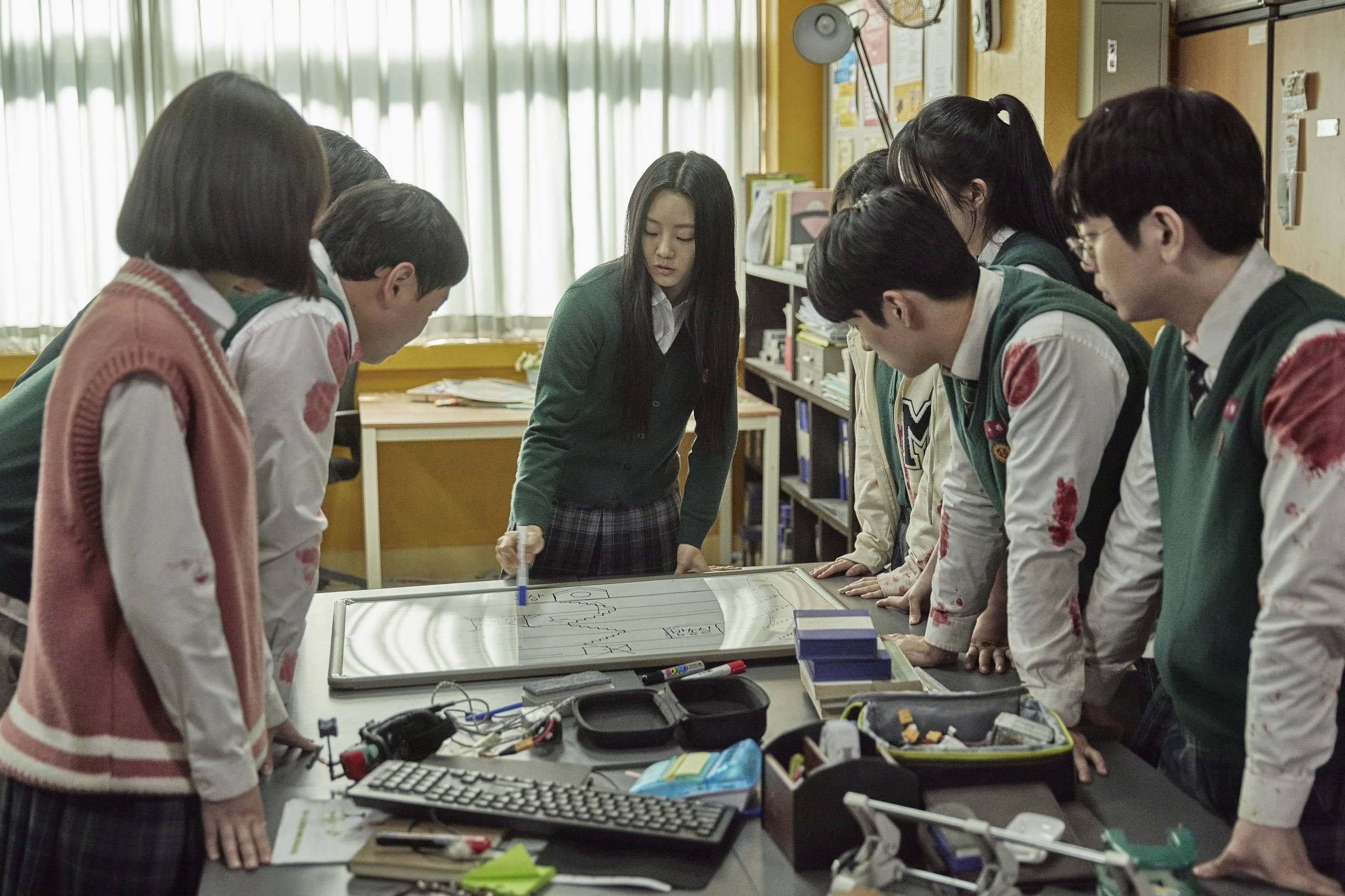 殭屍動圖 桌布 壁紙 — 身穿染血校服的學生擠在教室內一張雜亂的書桌旁，一名女孩正在白板上書寫。