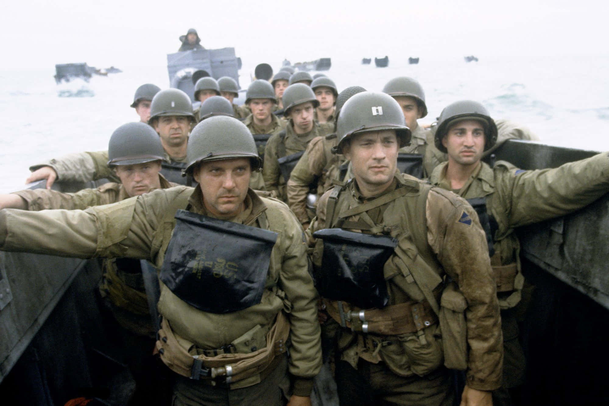 World War 2 桌布 壁紙 — 身穿橄欖綠制服和頭盔的士兵在登陸艇中緊張地站立，接近多雲天空下波濤洶湧的灰色大海。