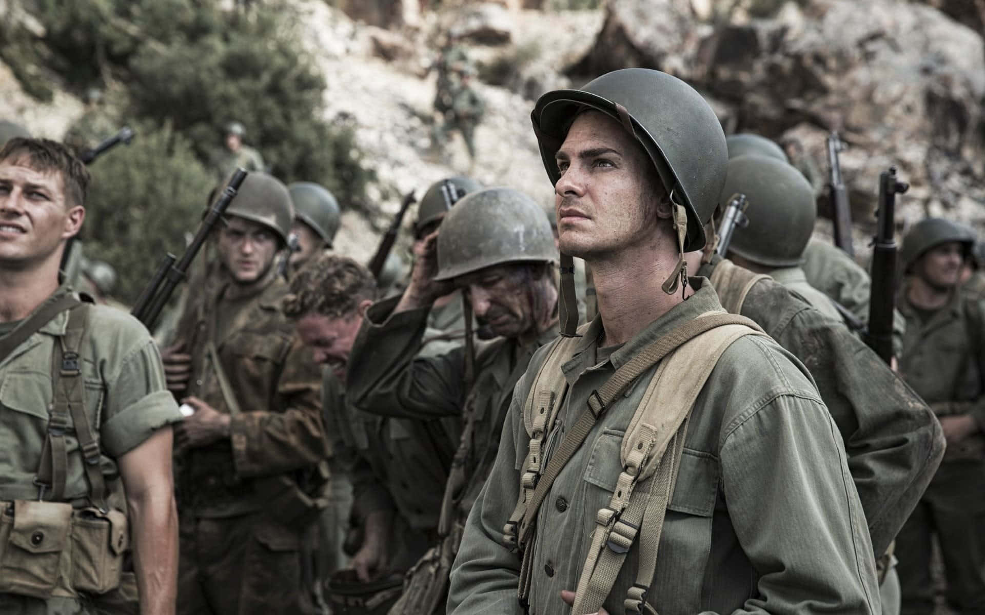 World War 2 桌布 壁紙 — 身穿橄欖綠制服和頭盔的士兵在自然日光下，以專注的表情站在岩石地形上。