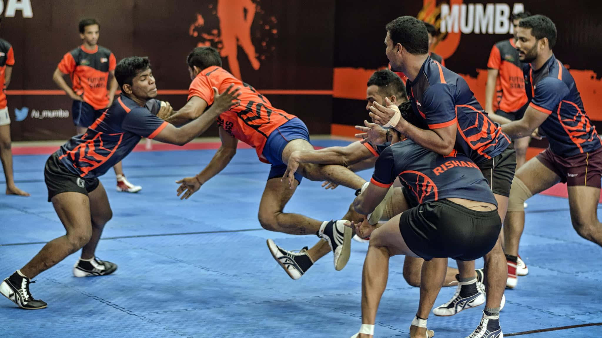 Kabaddi 桌布 壁紙 — 身穿橙色和海軍藍球衣的運動員在藍色墊子上激烈地扭打，進行一場競爭激烈的室內摔跤比賽。
