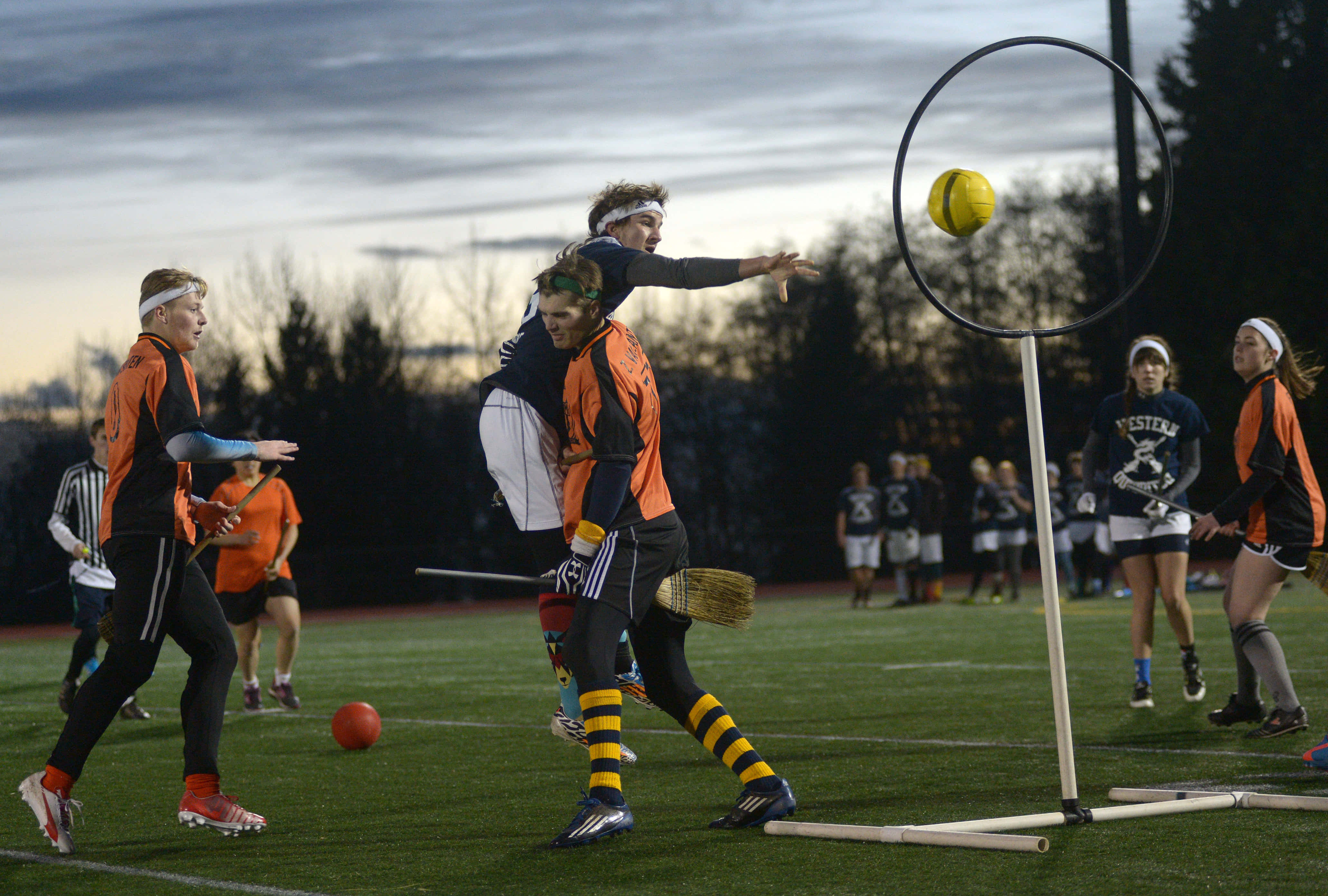 Quidditch 桌布 壁紙 — 身穿橙色和黑色制服的球員在黃昏的草地上追逐黃色的快浮穿過圓環。