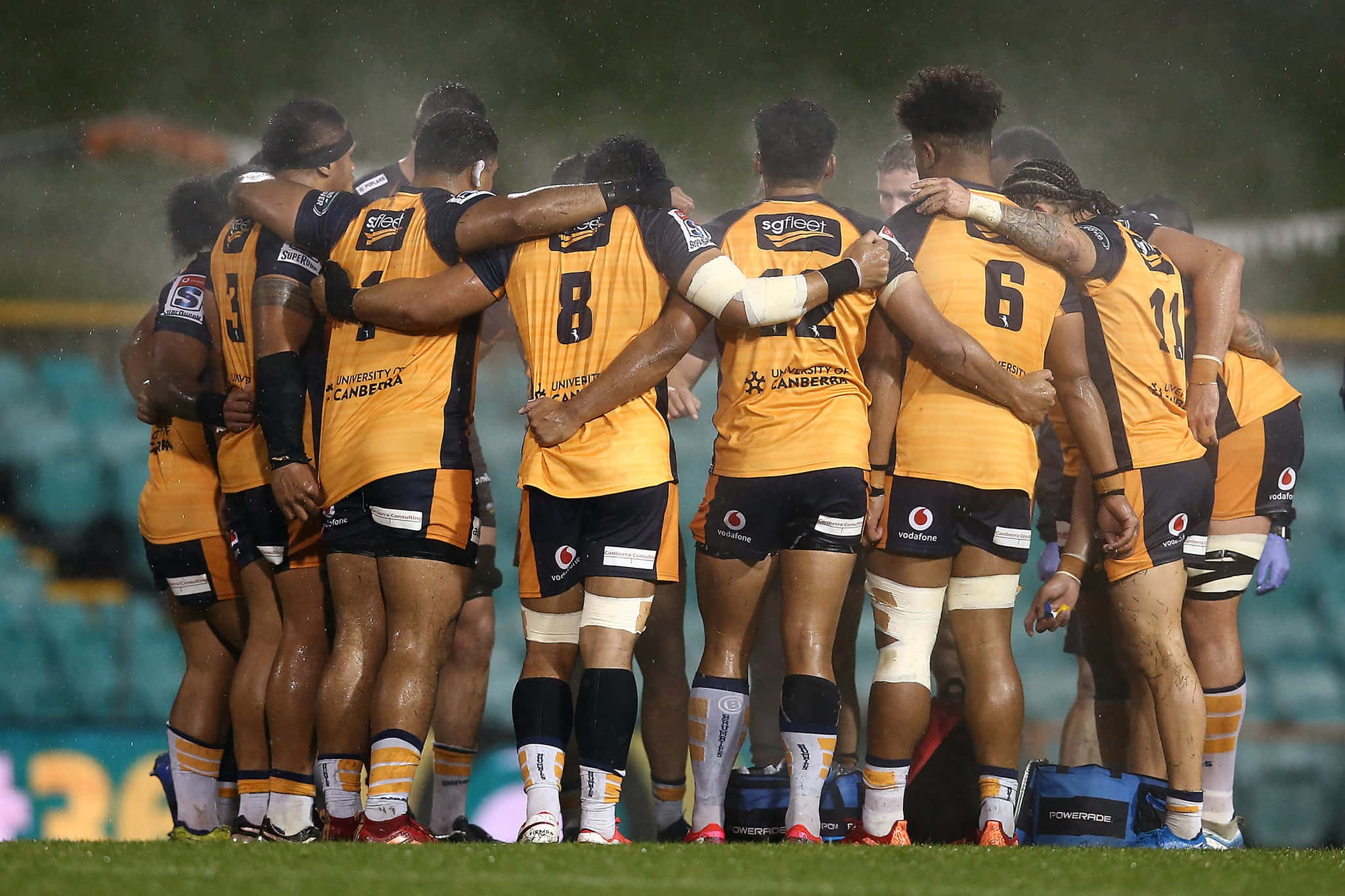 Brumbies 桌布 壁紙 — 身穿橙色和黑色球衣的橄欖球運動員在球場燈光下，在潮濕的場地上緊密地聚在一起。