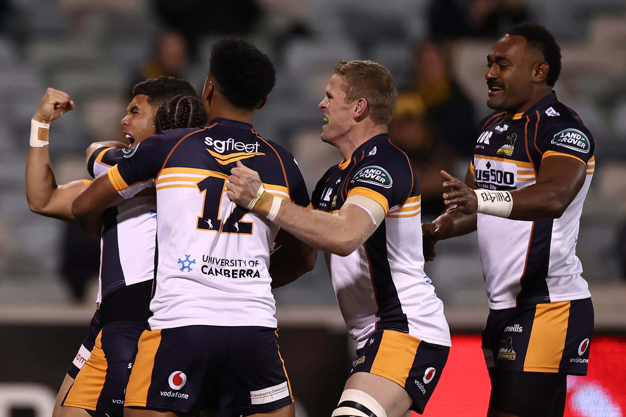 Brumbies 桌布 壁紙 — 身穿海軍藍和金色球衣的橄欖球聯賽球員在體育場燈光下舉起拳頭，帶著喜悅的表情慶祝達陣。