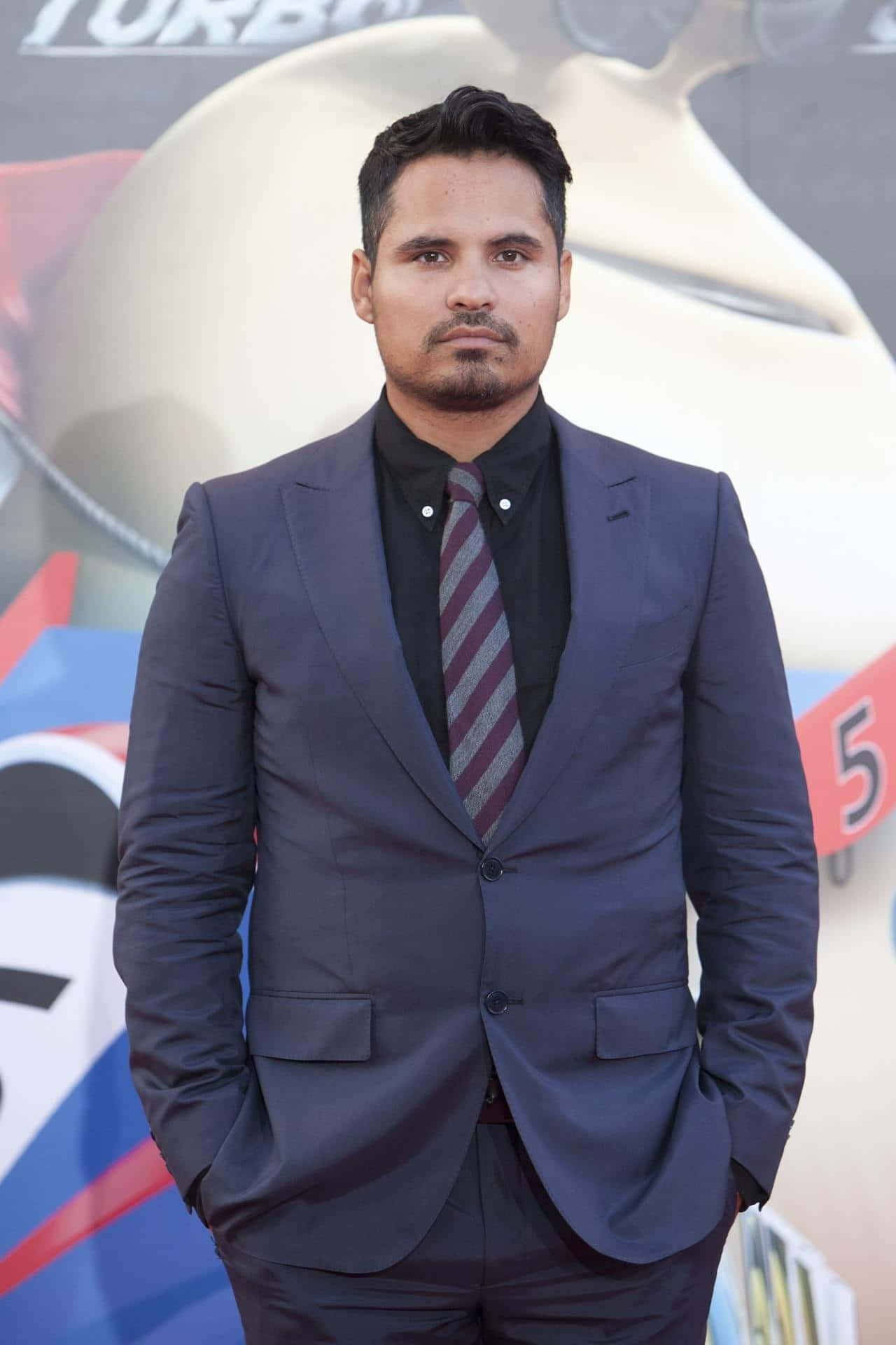 Michael Peña 桌布 壁紙 — 身穿海軍藍西裝和條紋領帶的男子自信地站在電影首映會上，雙手插在口袋裡。