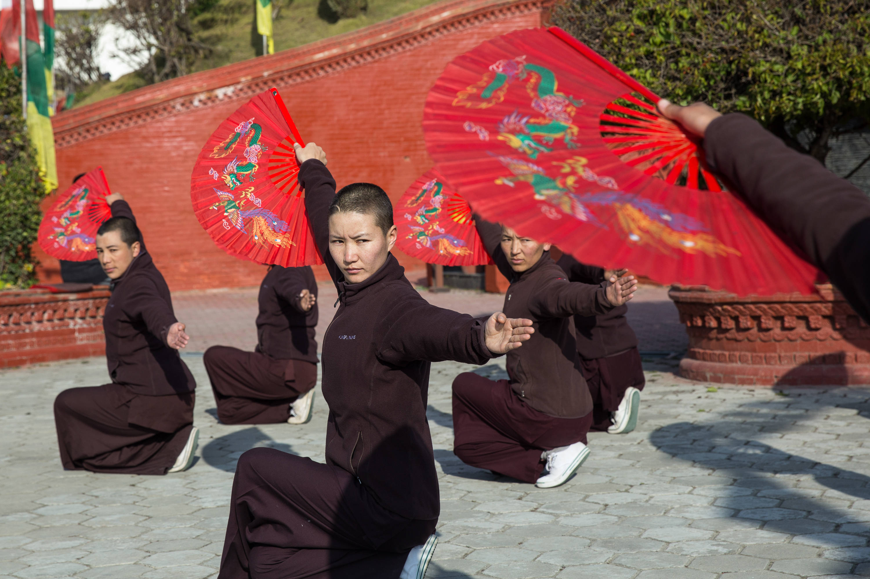 Exercise 桌布 壁紙 — 身穿深色長袍的僧侶在明亮的陽光下，以華麗的紅色扇子對著紅磚牆進行同步武術表演。