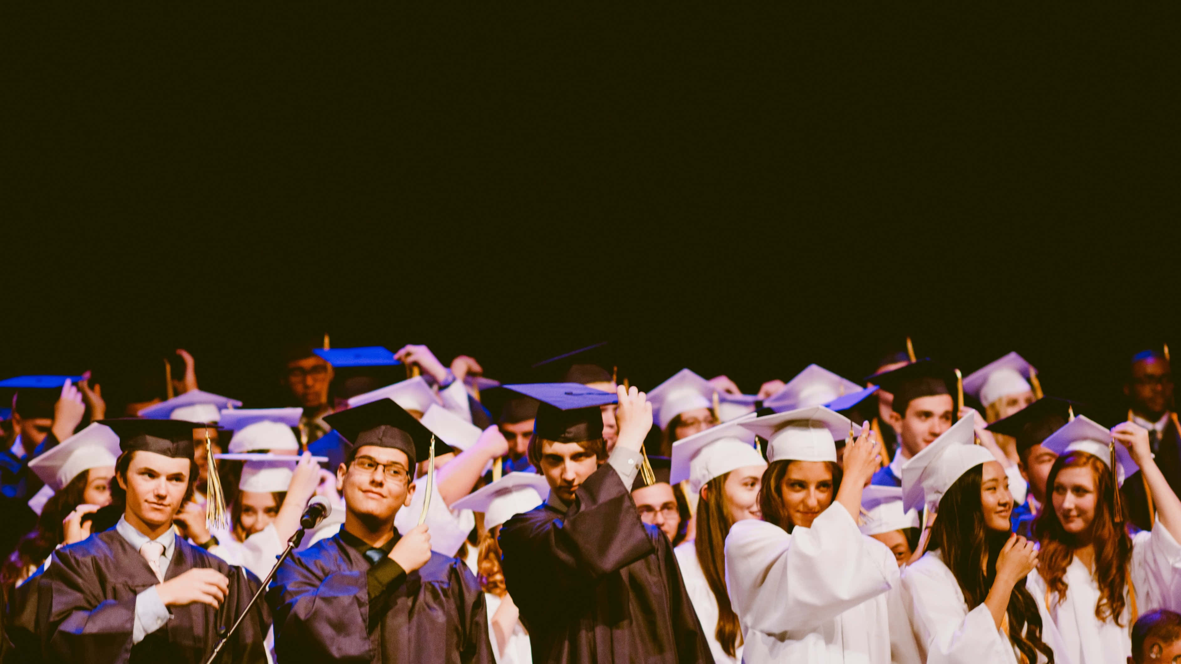 Graduation Ceremony 桌布 壁紙 — 身穿畢業袍和學士帽的畢業生在昏暗燈光下，於舞台上高舉雙手，帶著燦爛笑容歡樂慶祝。