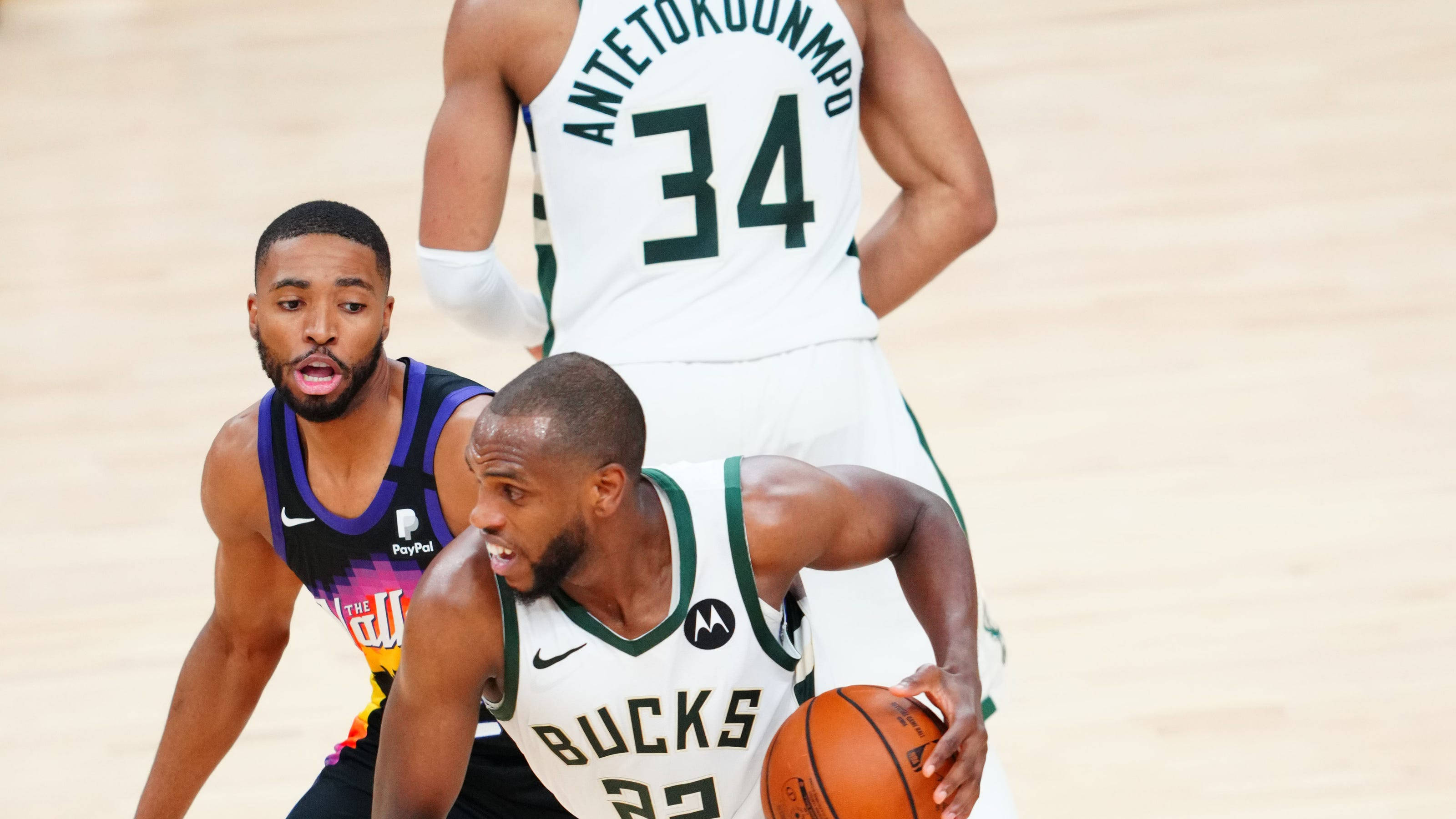 Khris Middleton 桌布 壁紙 — 身穿白色公鹿隊球衣的籃球運動員在激烈的比賽中運球，同時被身穿紫色球衣的對手防守。