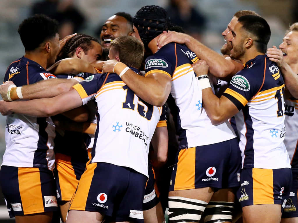 Brumbies 桌布 壁紙 — 身穿白色和橙色球衣的橄欖球運動員在球場燈光下緊密圍攏慶祝勝利。