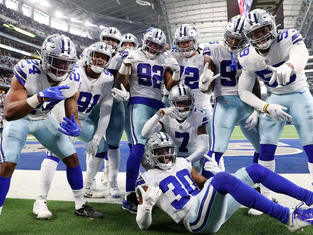 Dallas Cowboys Players 桌布 壁紙 — 身穿白色和藍色球衣的達拉斯牛仔隊美式足球員在體育場燈光下的綠色球場上一起慶祝。