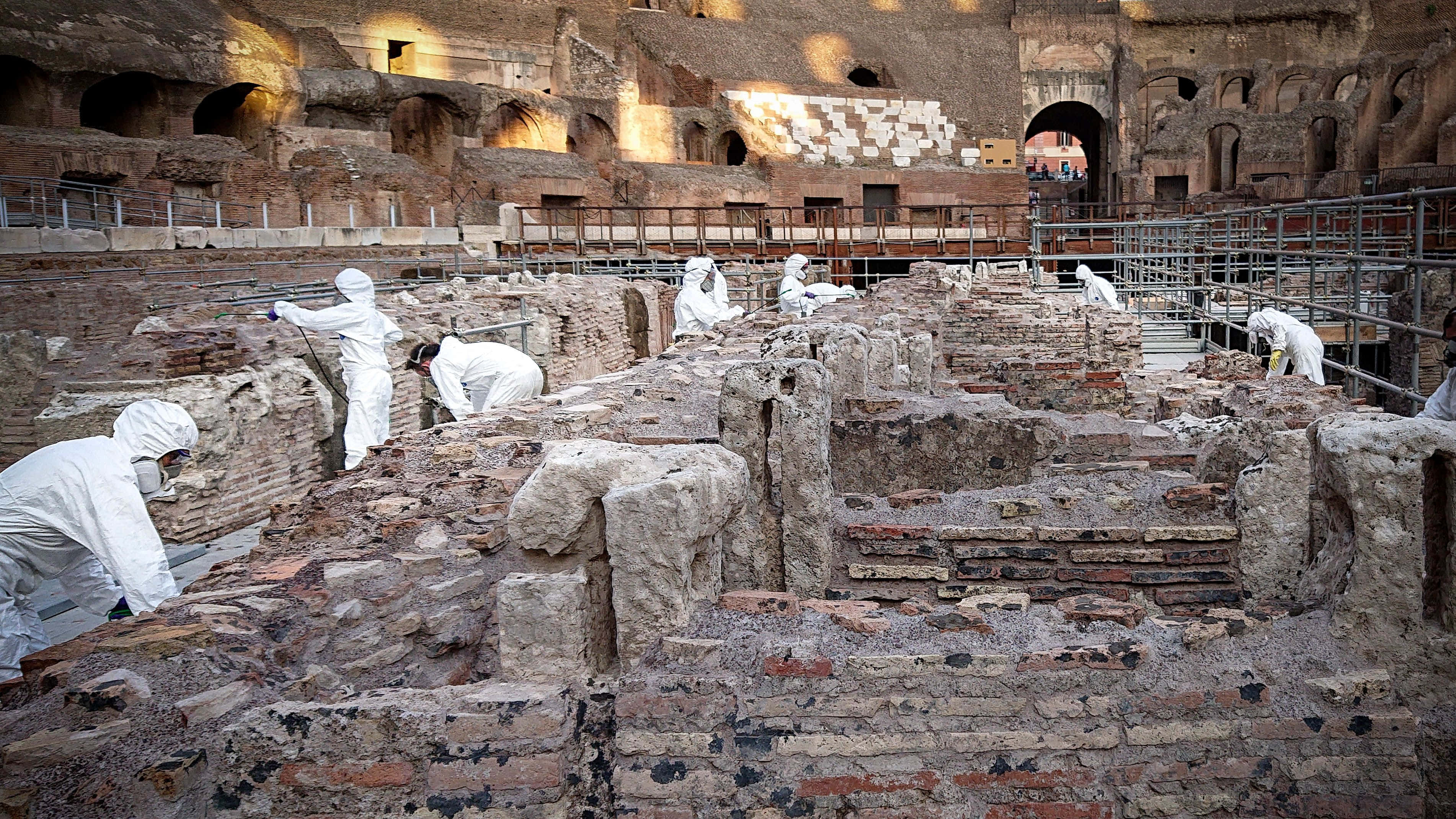 Roman Colosseum 桌布 壁紙 — 身穿白色套裝的考古學家在溫暖的陽光下，在羅馬競技場內仔細挖掘古羅馬遺址。