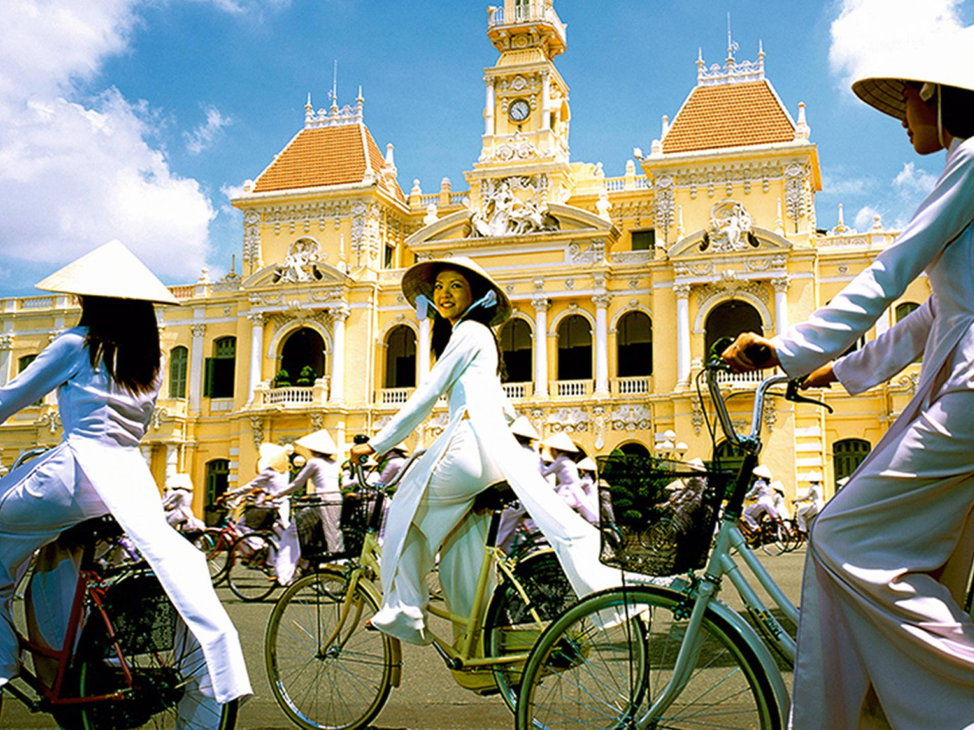 Ho Chi Minh City 桌布 壁紙 — 身穿白色奧黛和錐形帽的女性騎著腳踏車，在明亮的藍天下經過一座宏偉的黃色殖民建築。