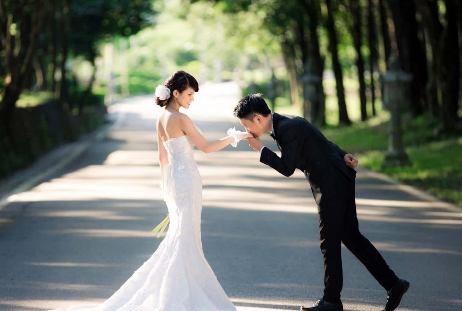 Wedding Couple 桌布 壁紙 — 身穿白色婚紗的新娘和身穿黑色西裝的新郎在陽光普照的林蔭小徑上分享溫馨時刻。